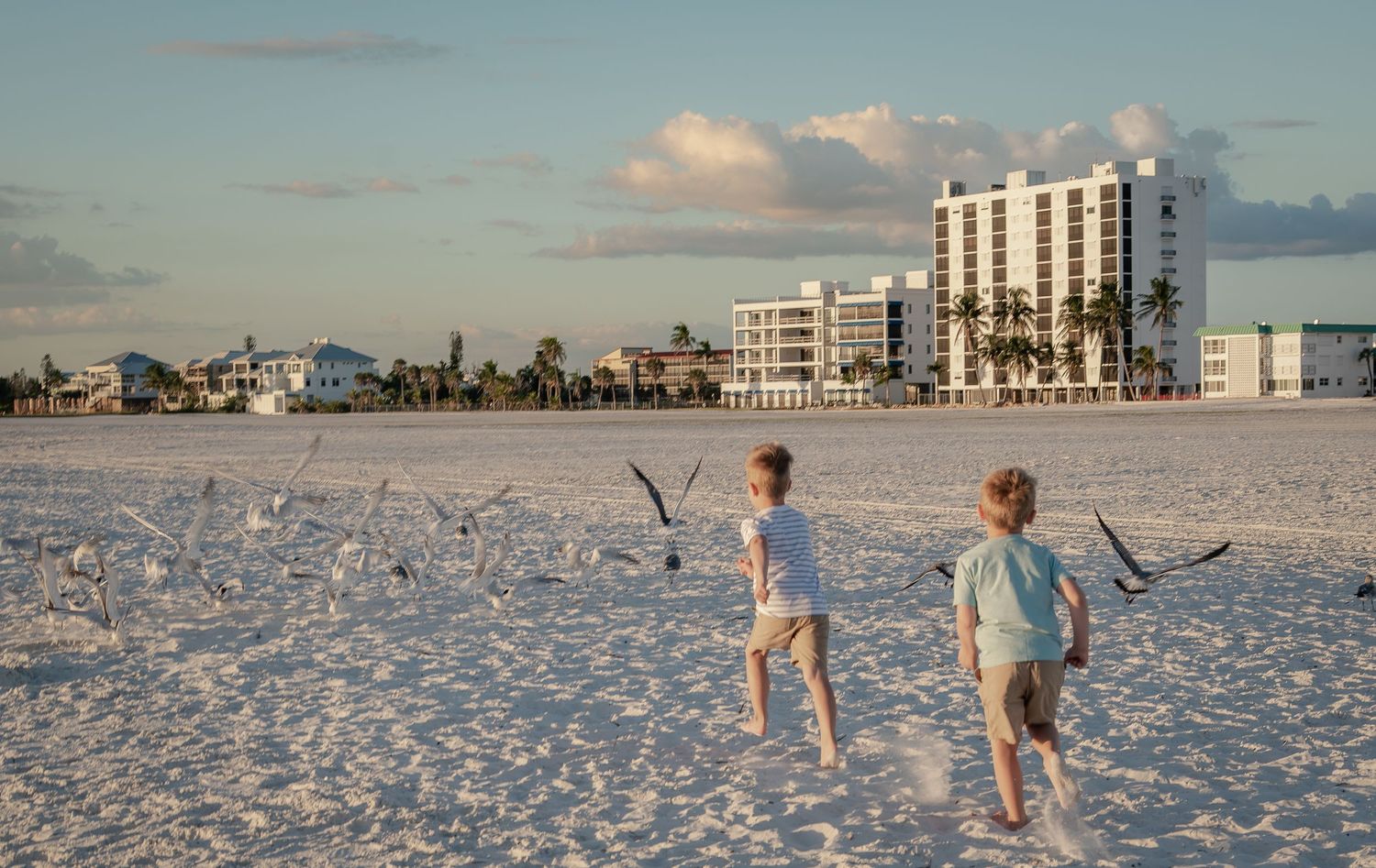 Two young boys running barefoot on white sand beach toward seagulls with buildings in background.
