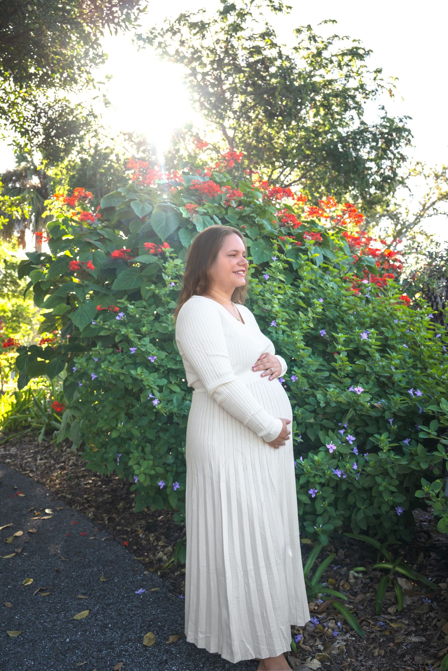 Pregnant woman in white pleated dress standing in garden with colorful flowers in bright sunlight.