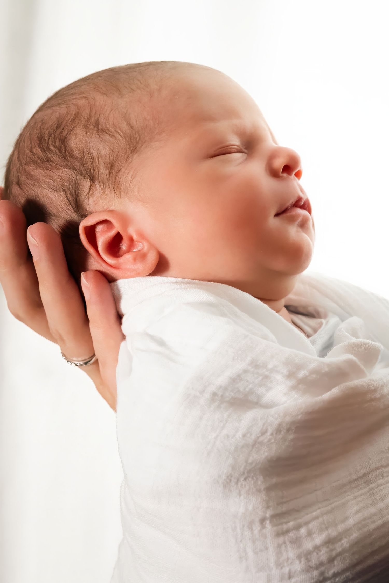 Peaceful newborn baby swaddled in white muslin cloth, sleeping while cradled in a parent's gentle hands.