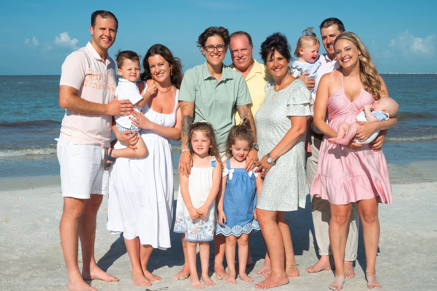 Large family group posing together on a sunny beach, including adults, children, and babies, smiling for a portrait.