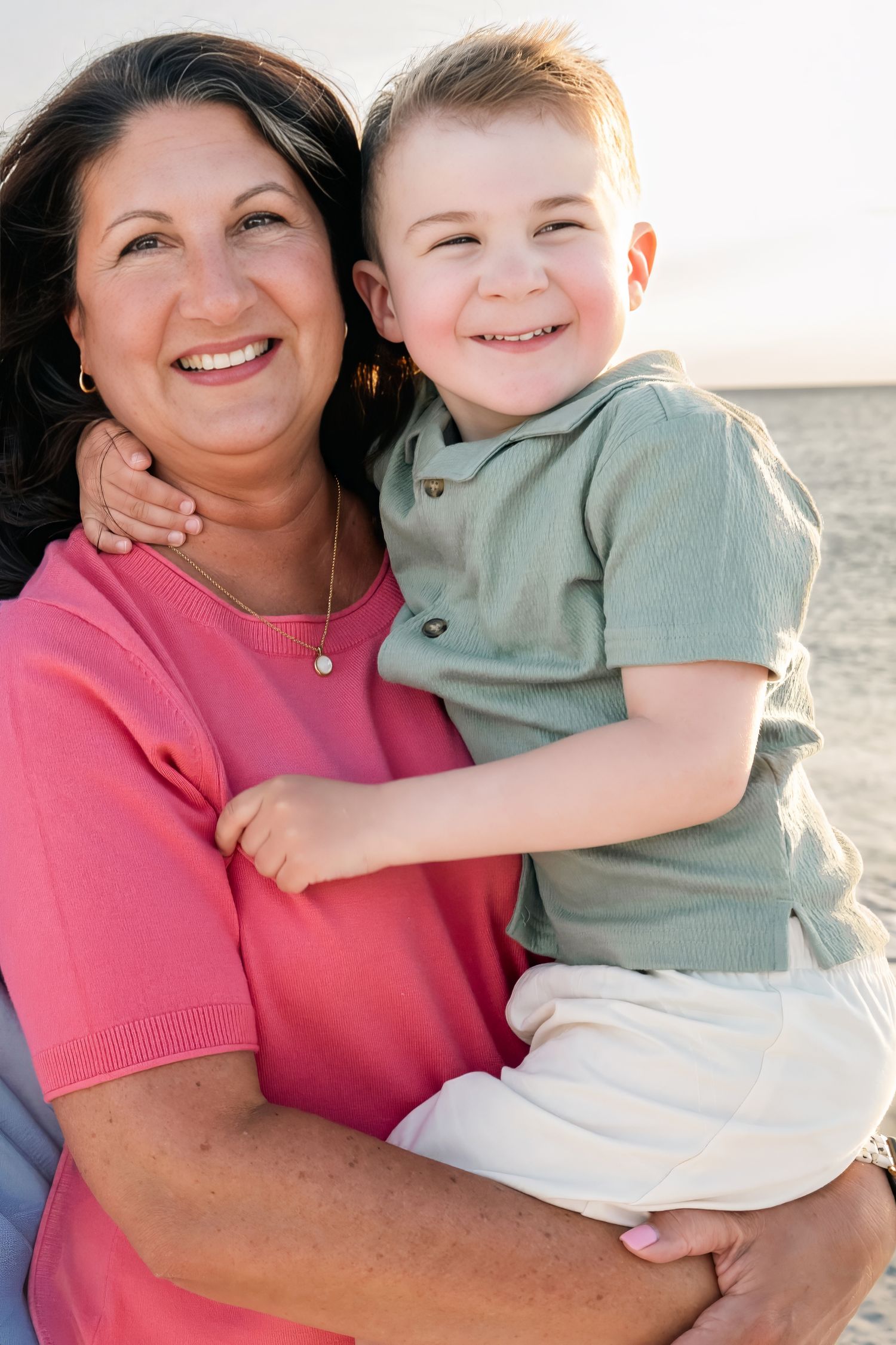 A smiling woman in a pink shirt holds a laughing young boy near the beach in warm sunlight.