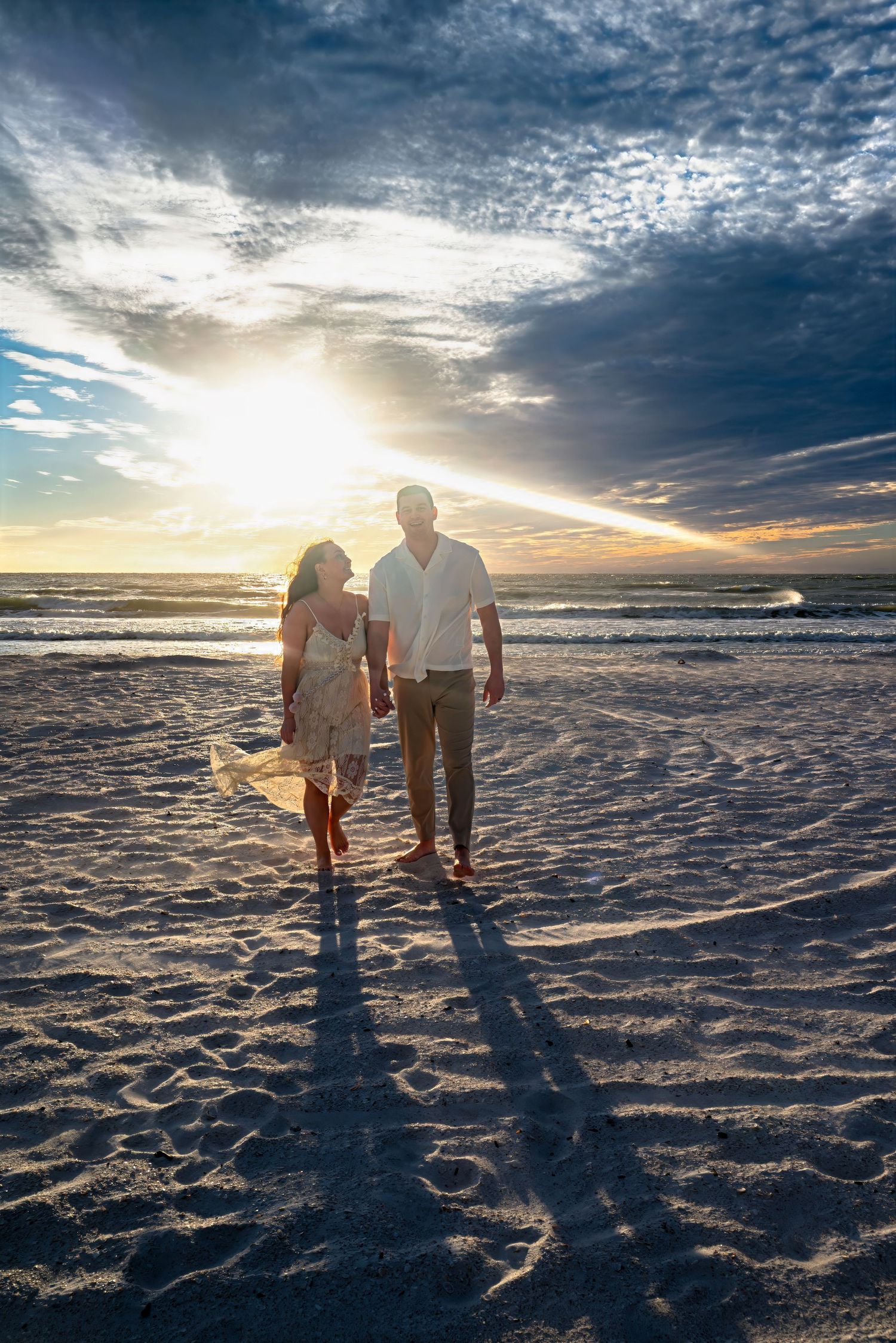 Couple holding hands walking on a sandy beach at sunset with dramatic cloudy sky.