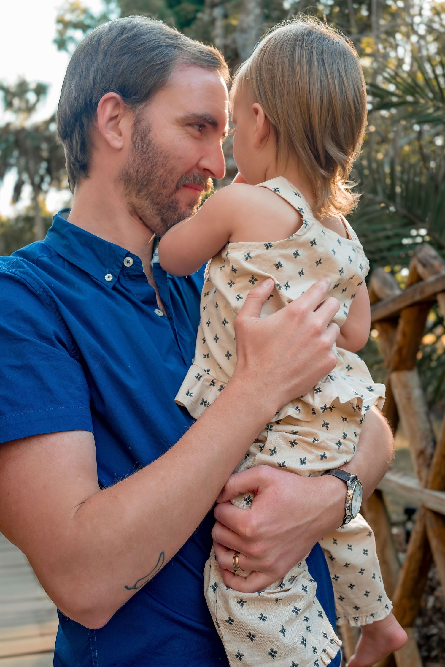 A bearded man in a blue shirt tenderly holds a toddler in a floral romper outdoors.