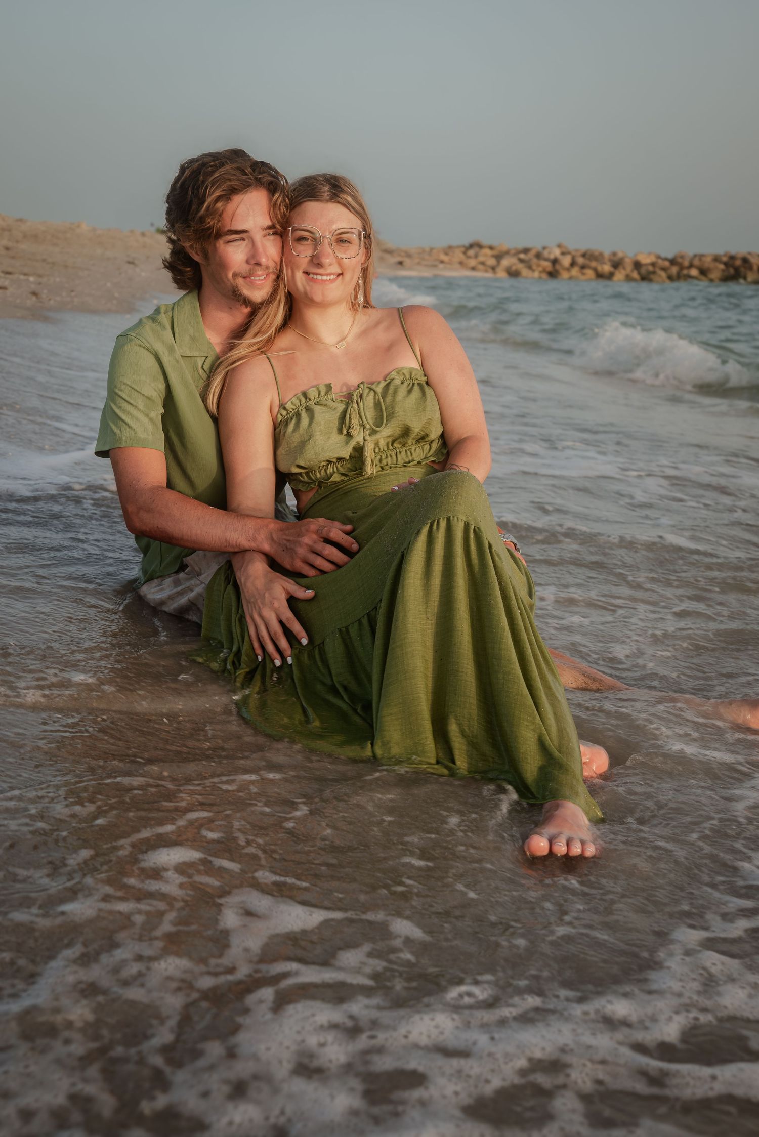 Happy couple sitting together in shallow ocean waves on beach at sunset, smiling and embracing each other.