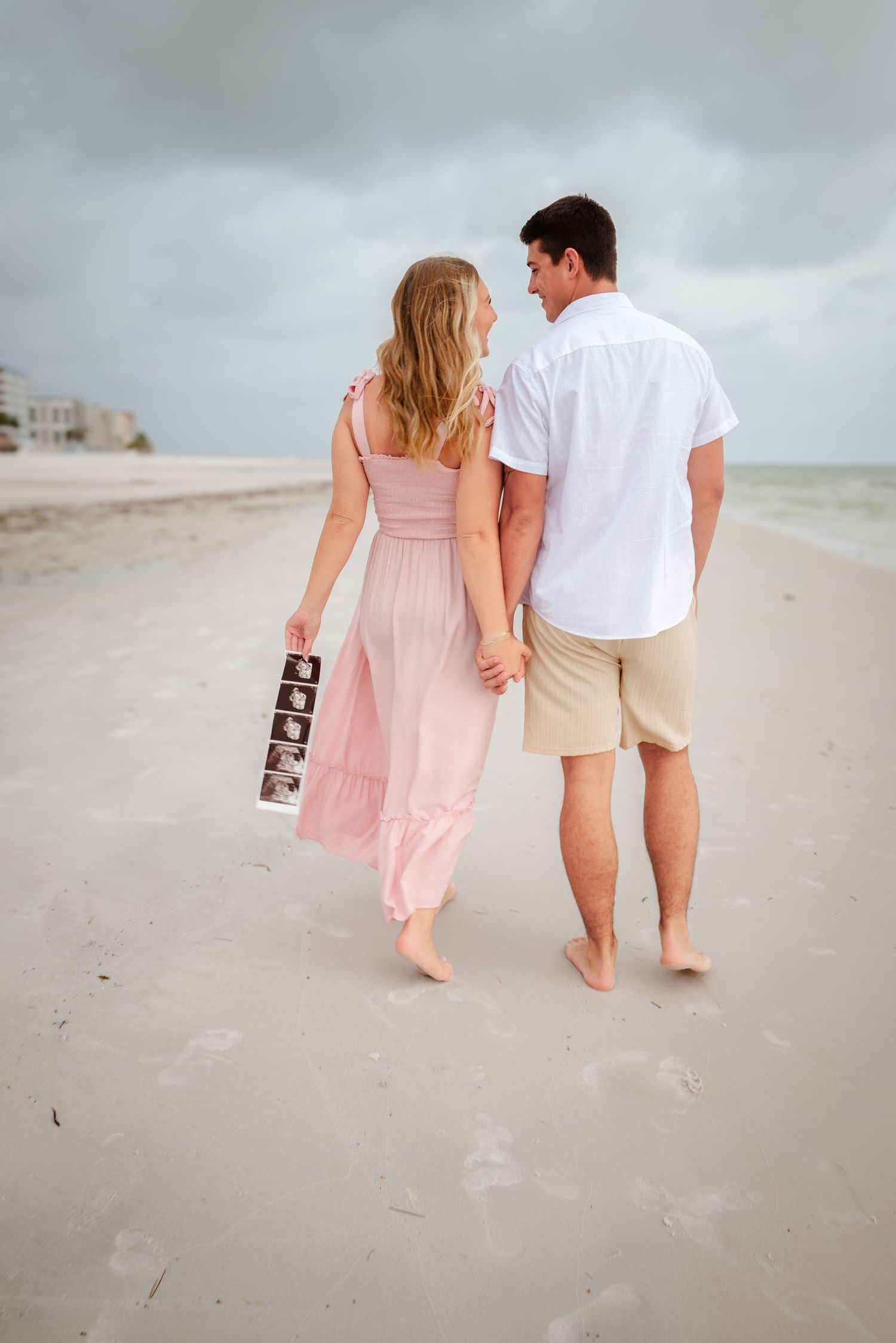 Couple walking on the beach holding hands and ultrasound photos on a cloudy day.