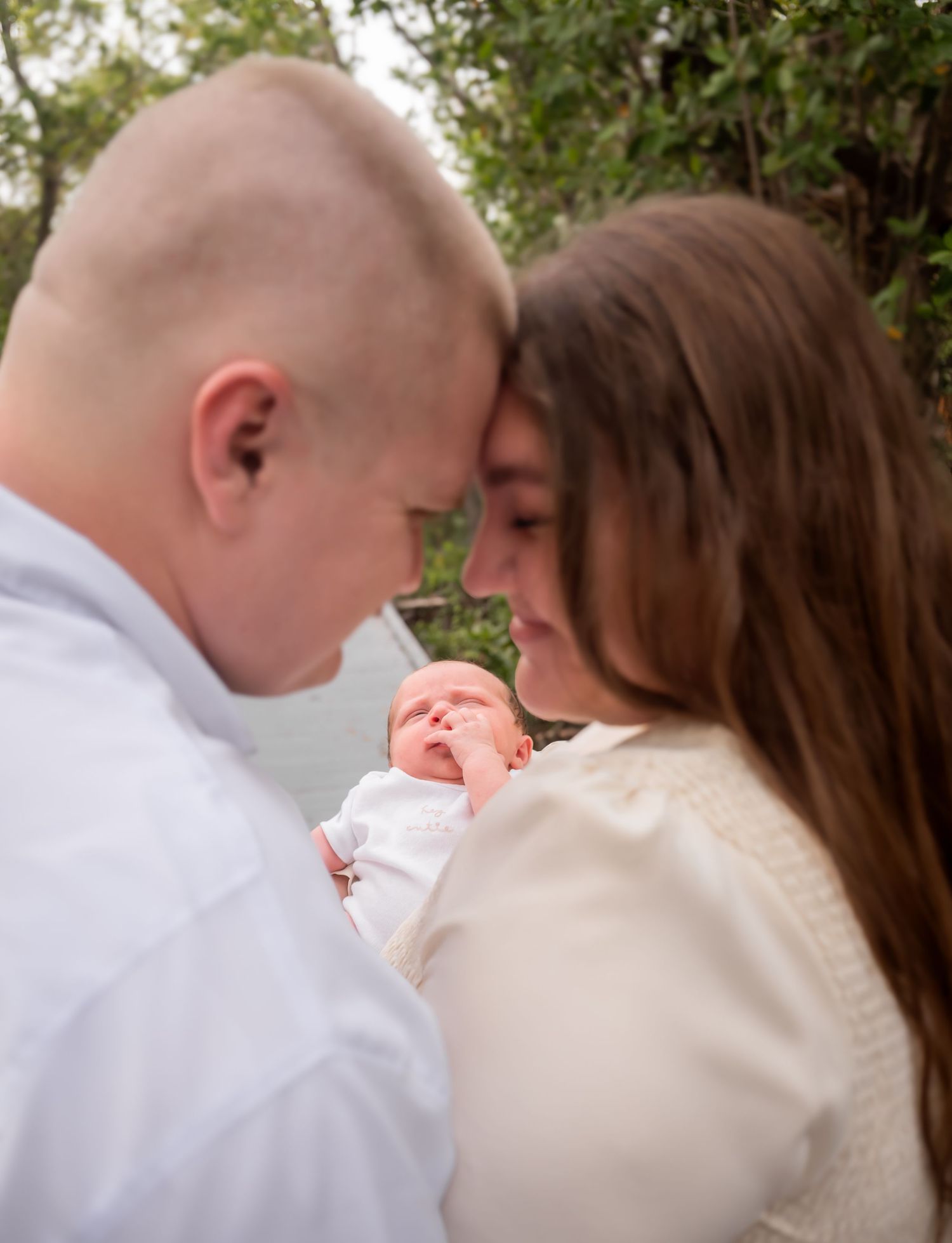 Couple tenderly touches foreheads while holding their sleeping newborn baby outdoors.