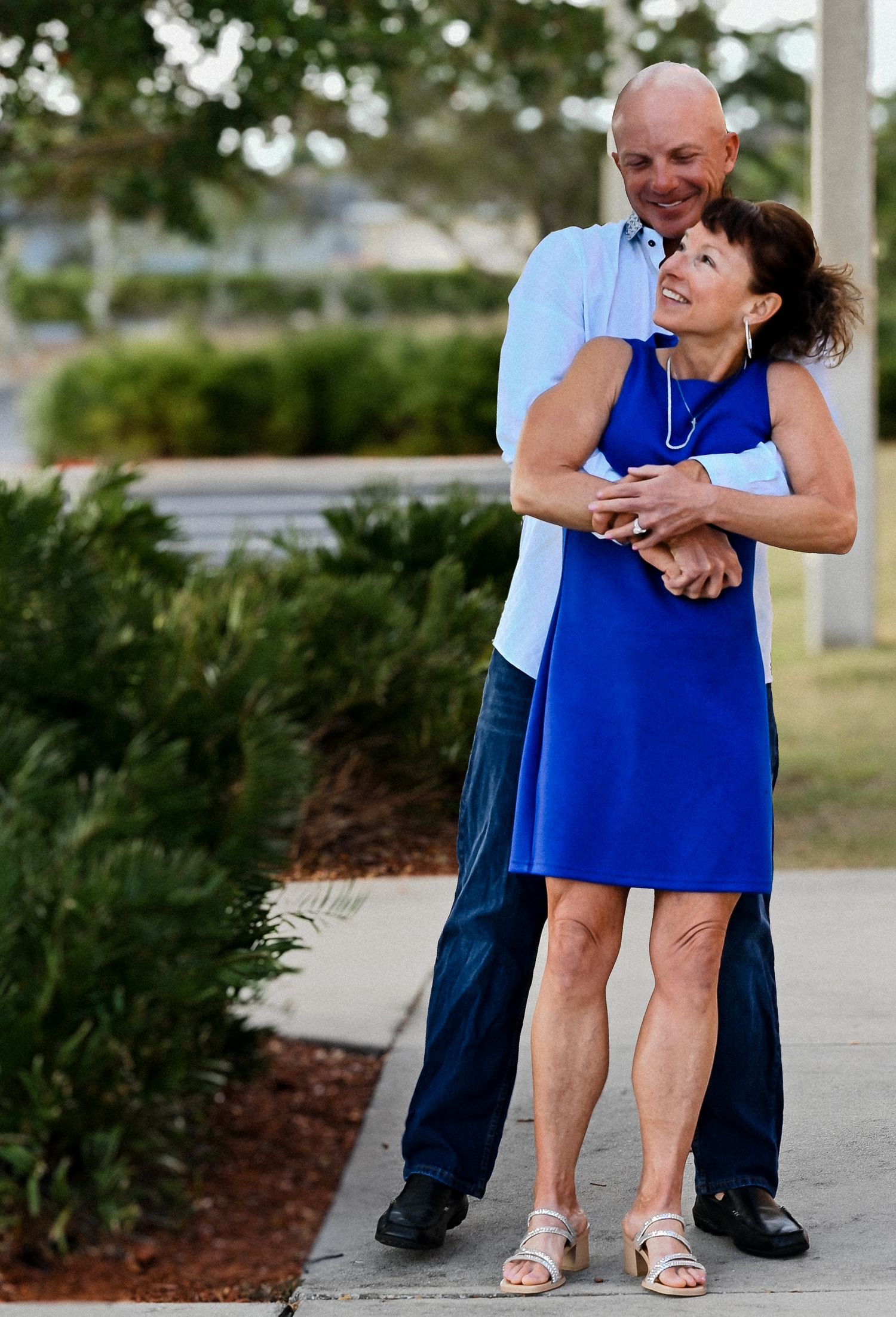 Bald man embraces woman in royal blue dress from behind on sunny outdoor walkway.