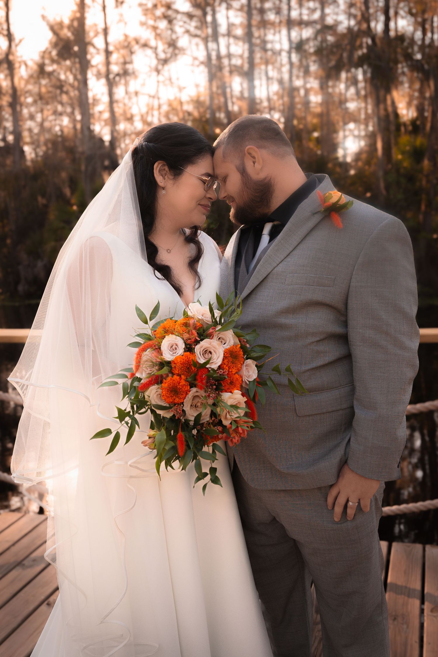 Bride and groom touch foreheads tenderly on wooden dock at golden hour sunset.