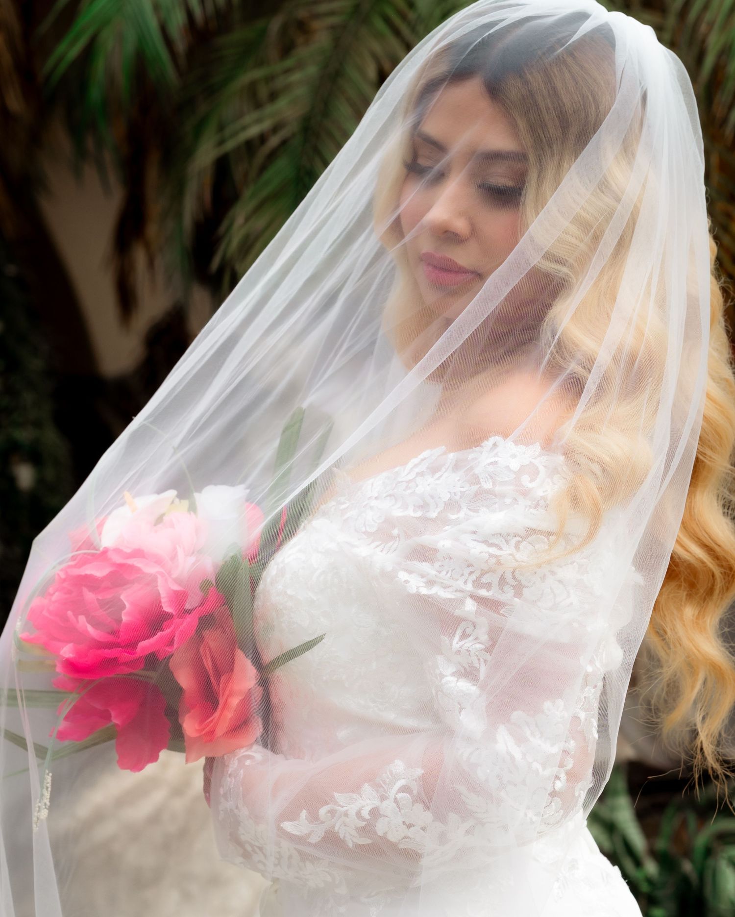 Bride in lace wedding dress and sheer veil holding pink peony bouquet, looking down gracefully.