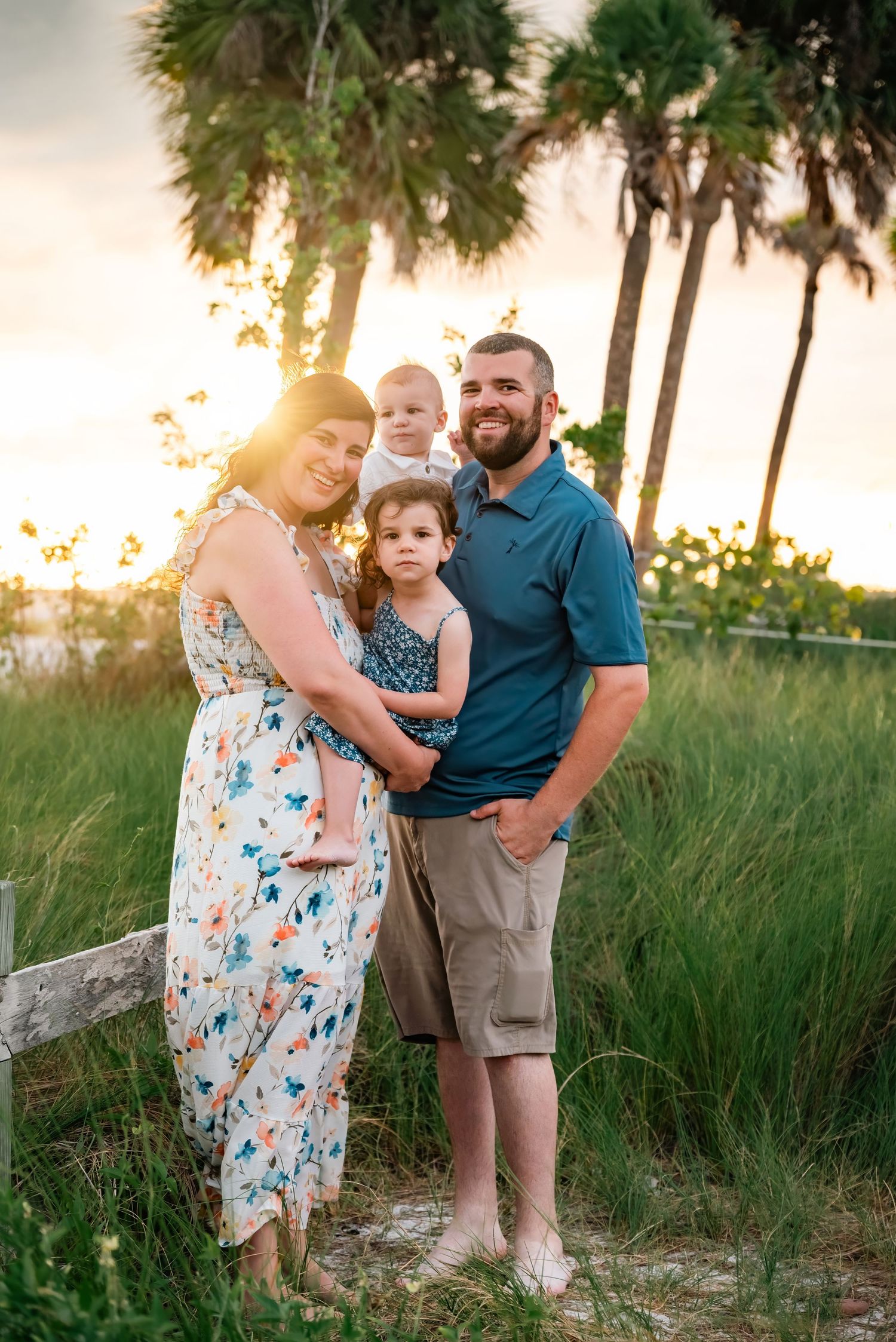 Family of four poses at sunset near palm trees and lush greenery, mom in floral dress, dad in teal polo holding toddler.