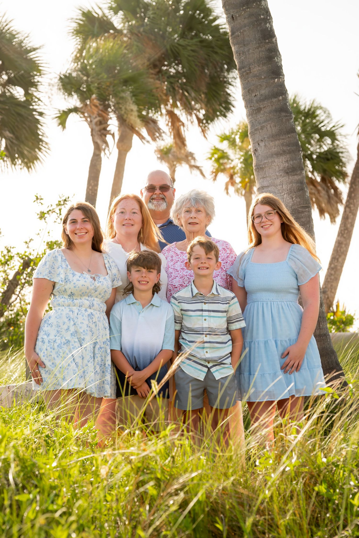 Multi-generational family of seven smiling outdoors among palm trees and green grass in a sunny coastal setting.