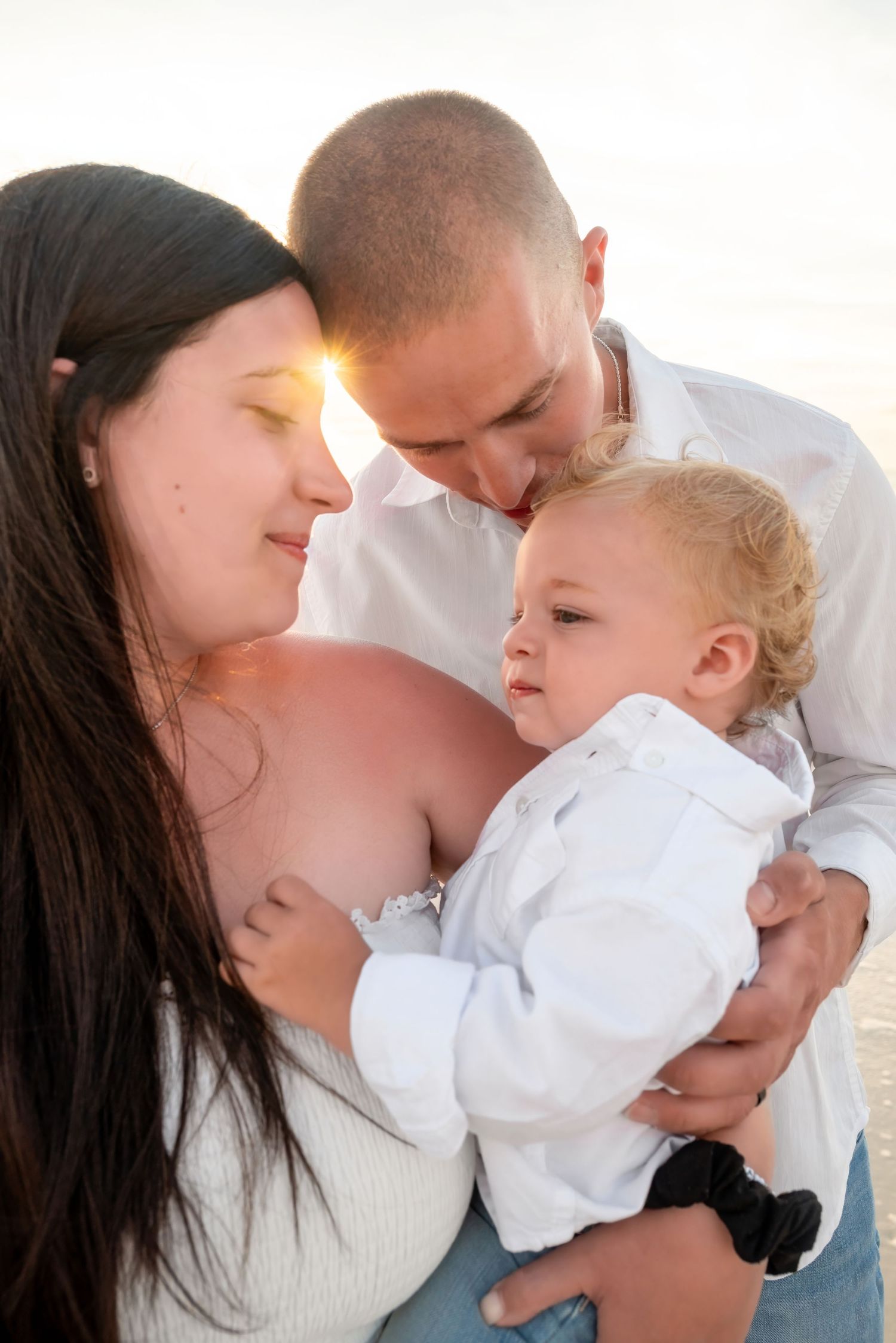 A loving family with a pregnant mother, father, and baby dressed in white sharing a tender moment on a beach.