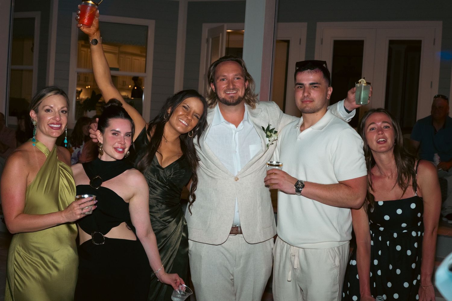 Wedding reception group photo with groom in tan suit celebrating with guests raising drinks indoors.