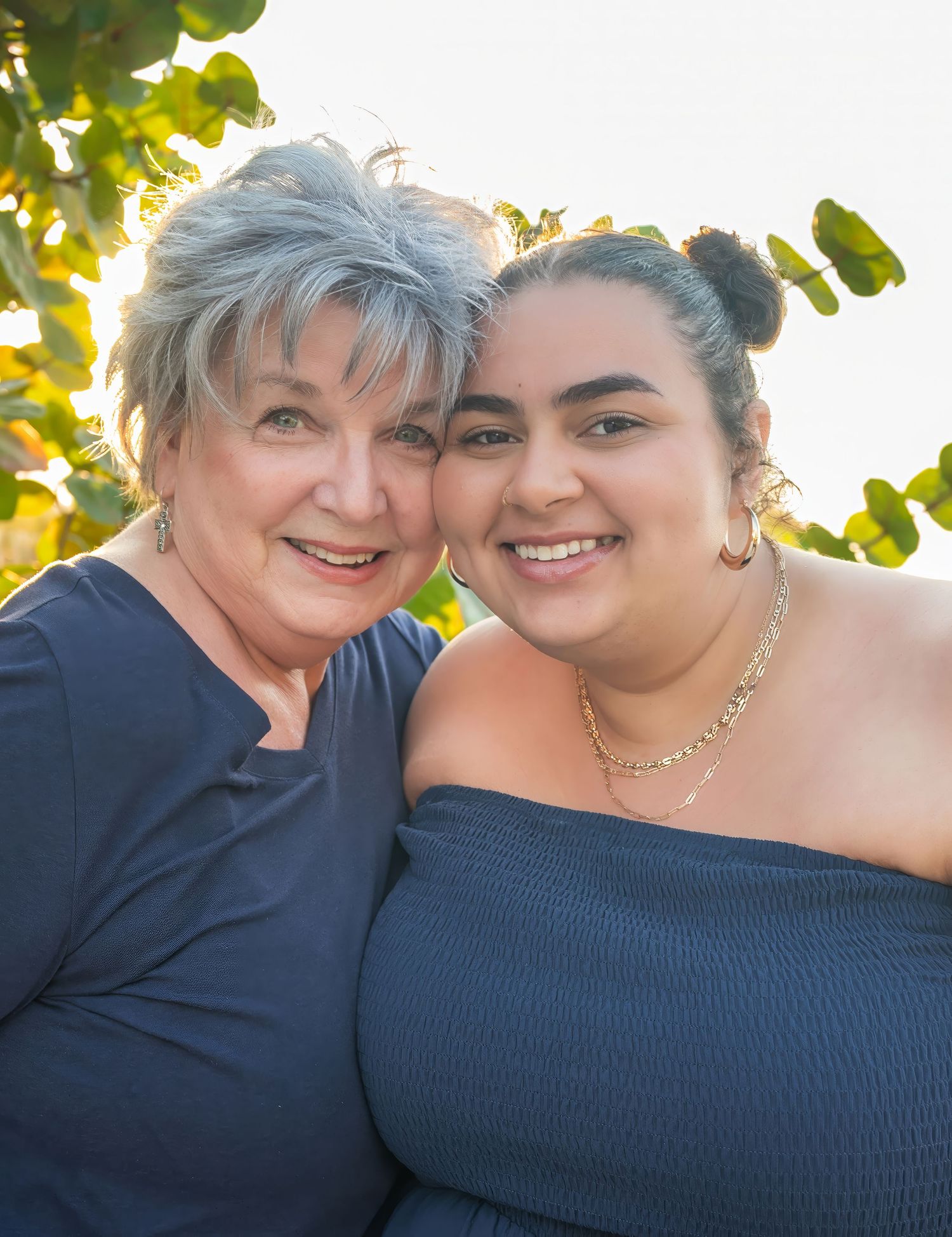 Two women, one with silver hair and one younger, smiling warmly together outdoors in golden light.