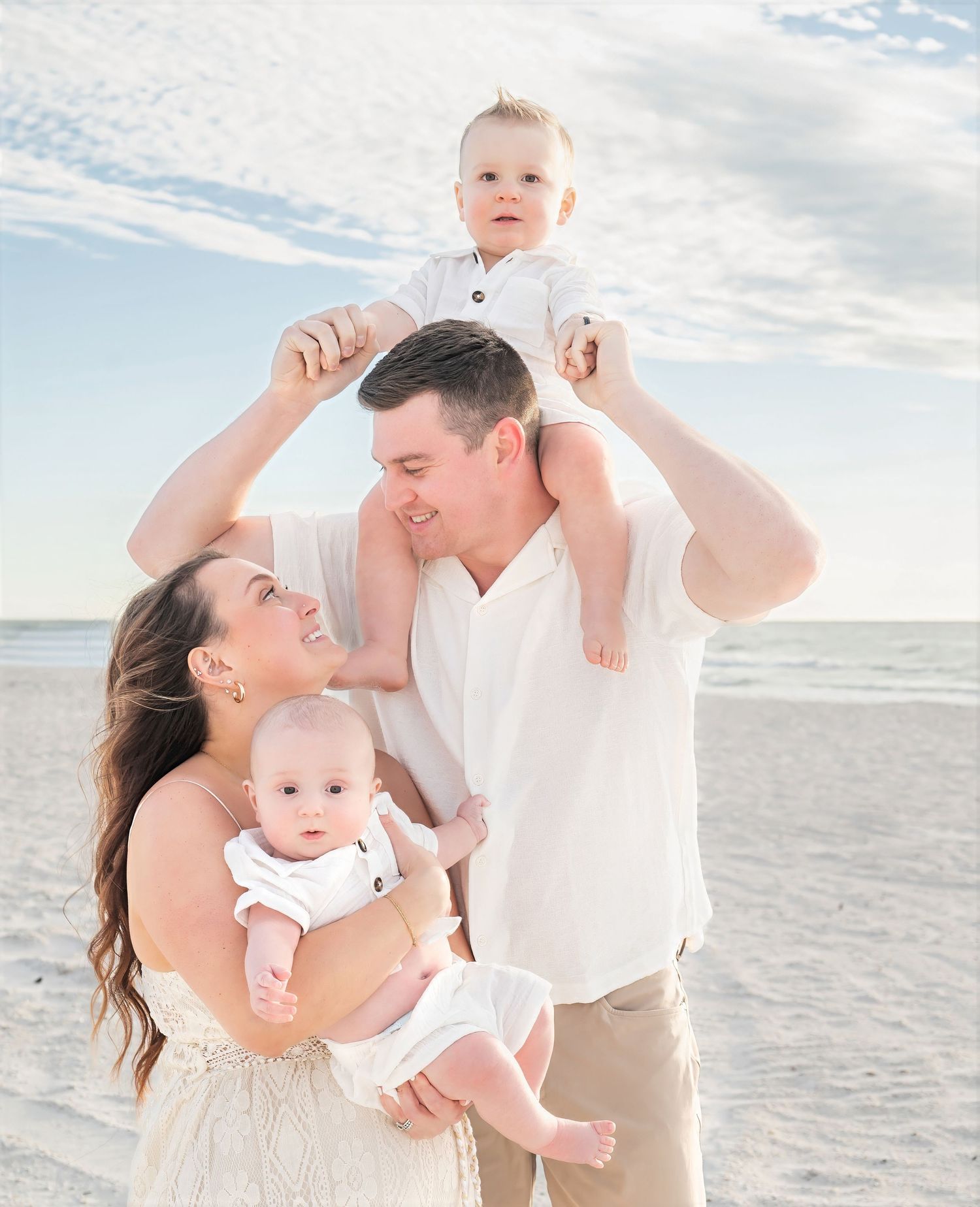 A couple on a beach holds two young babies while smiling at each other.