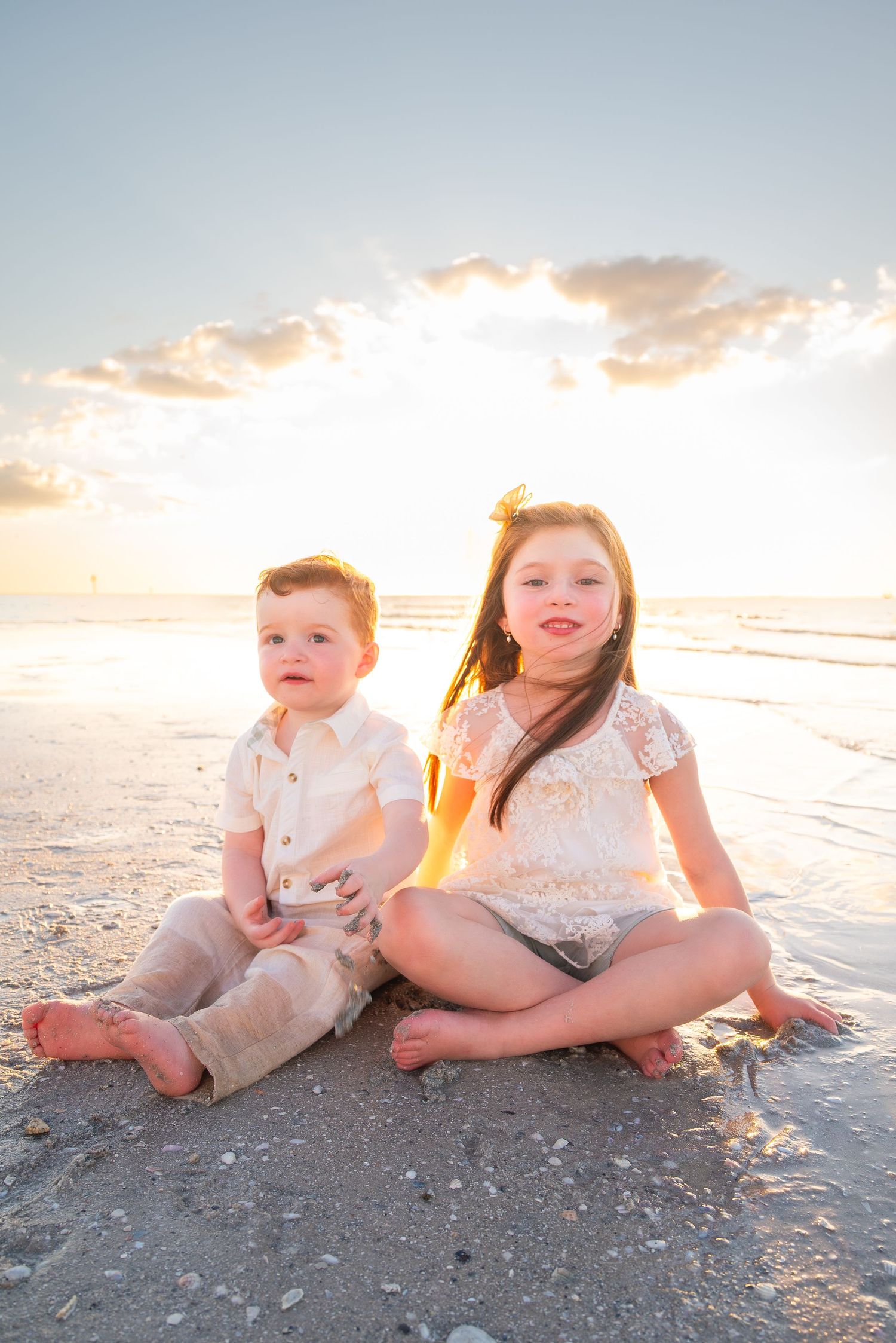 Two young children sit together on a sandy beach at golden sunset, dressed in white outfits.