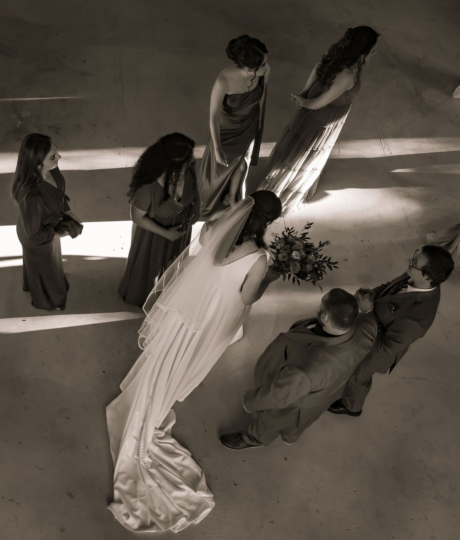 Aerial black and white view of bride in white gown surrounded by wedding party indoors.