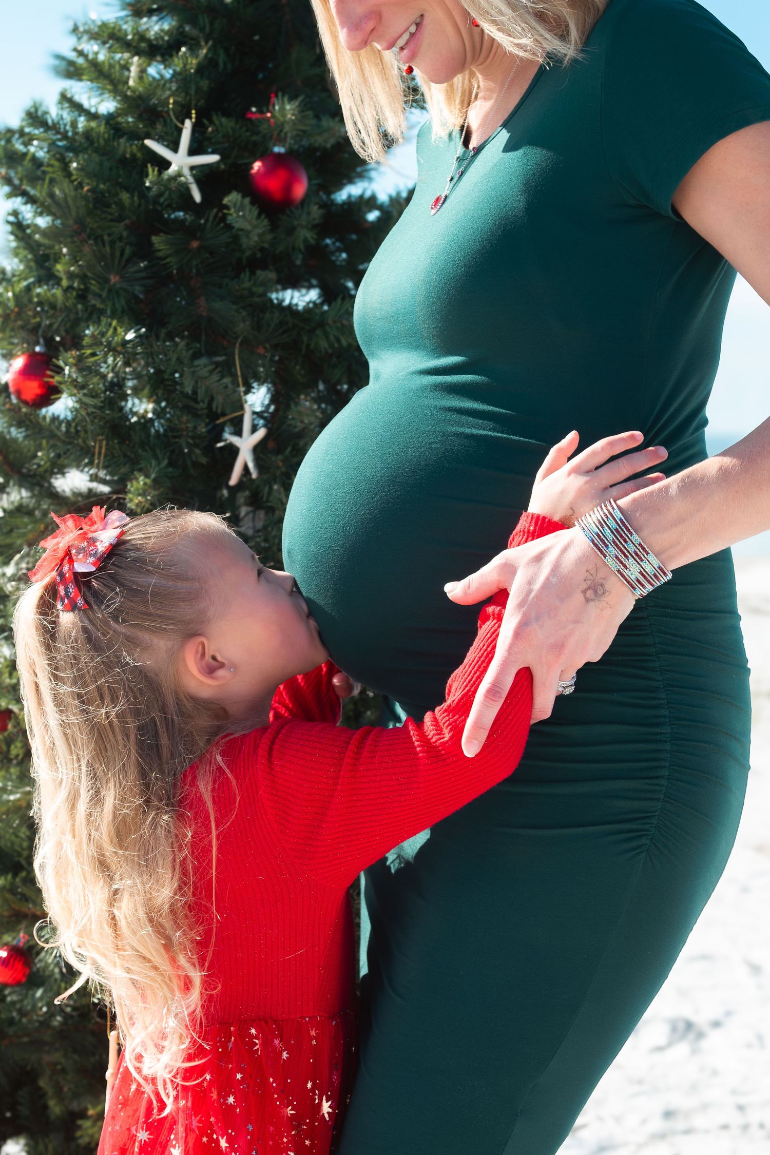 Young girl kissing pregnant mothers belly in front of a beach Christmas tree.