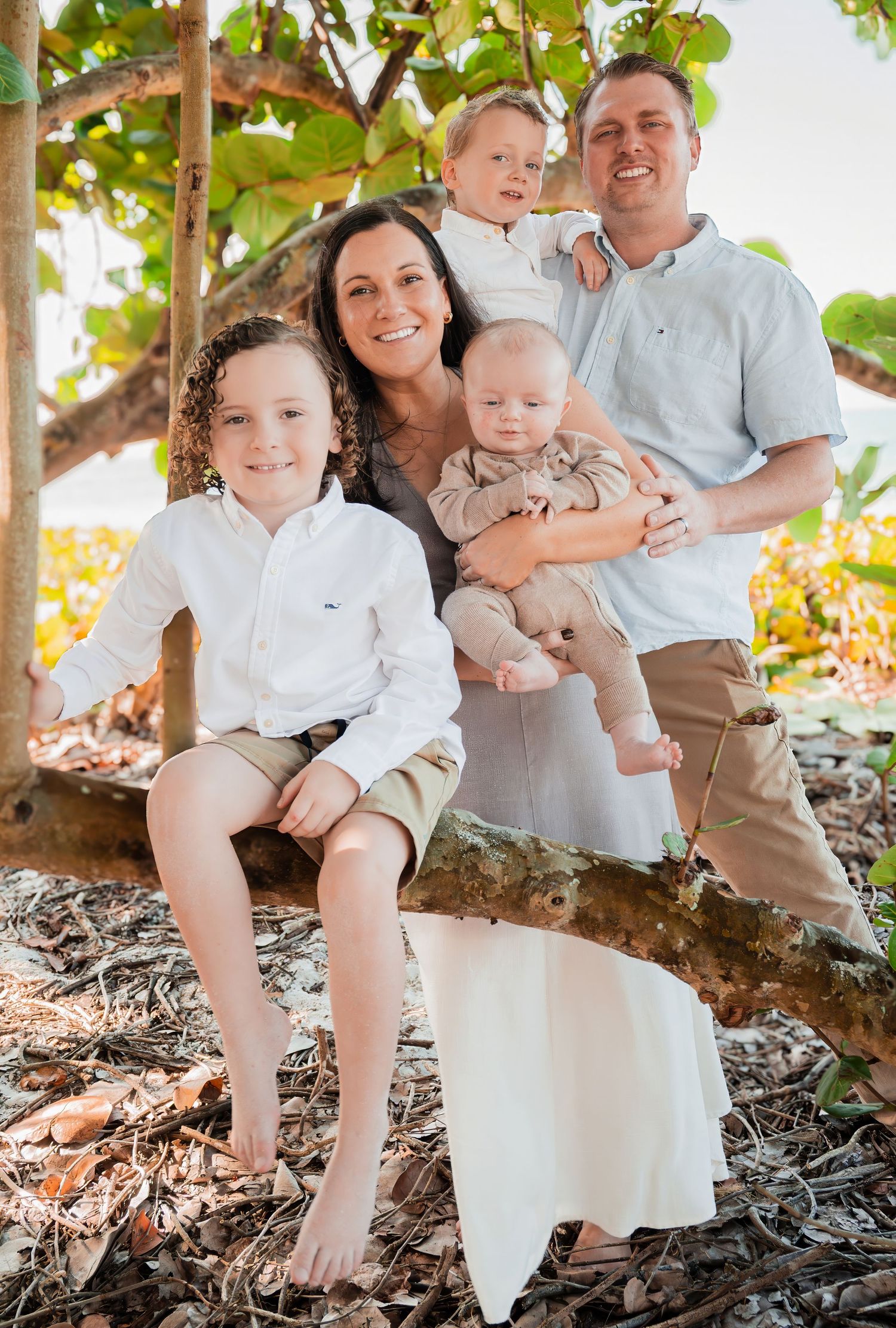 Happy family of five posing under a tropical tree in coordinated neutral outfits outdoors.