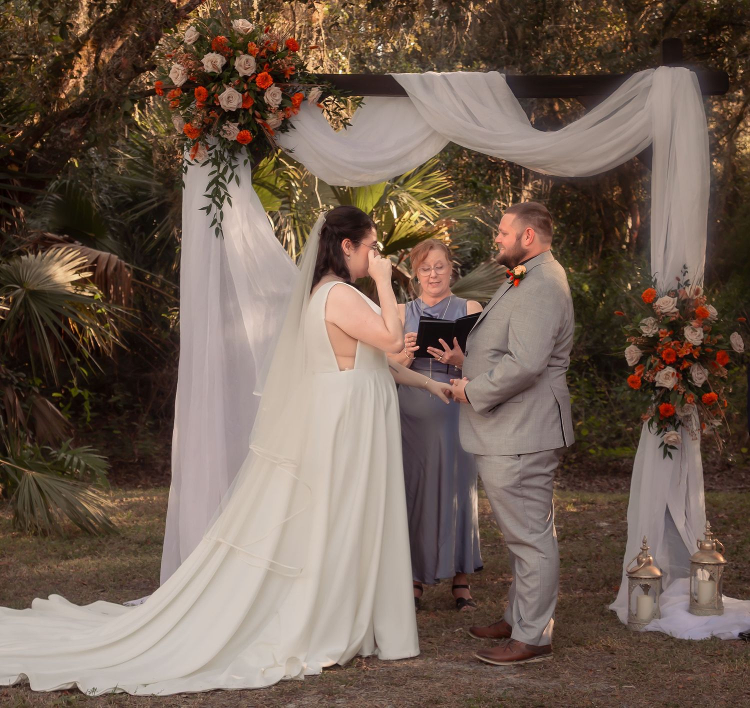 Bride and groom exchange vows beneath a white draped floral arch at an outdoor woodland wedding.