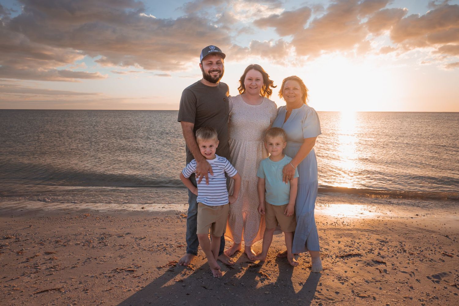Family of five posing together on a beach at sunset with golden clouds reflecting over calm ocean waters.