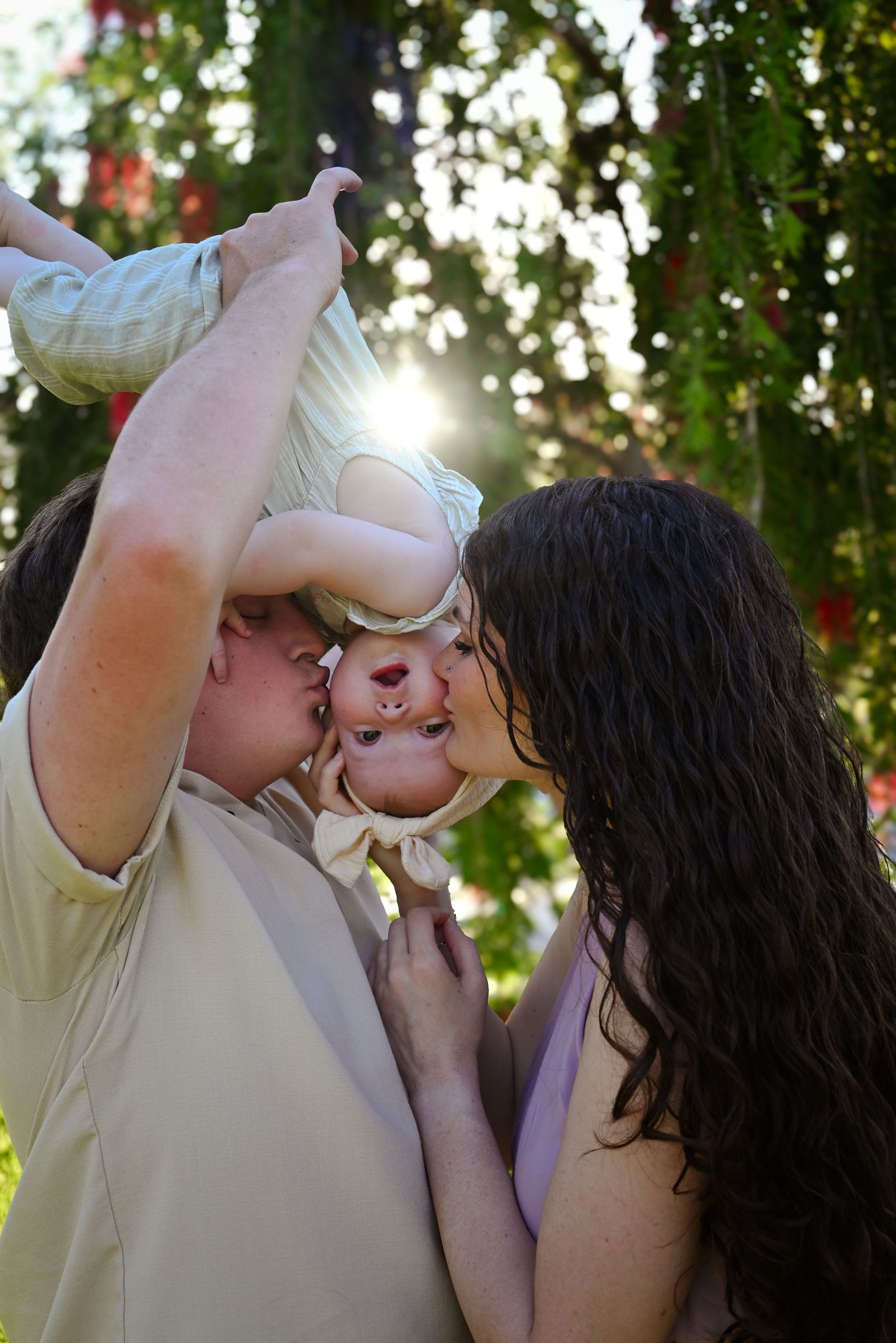 Parents kissing their baby held upside down in sunny garden.