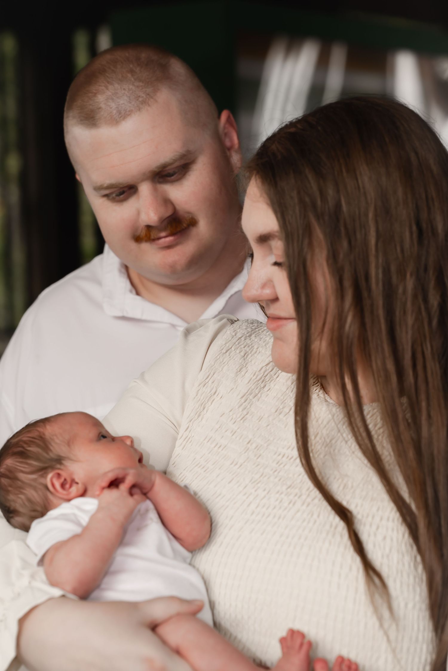 New parents in white outfits lovingly gazing at their newborn baby together outdoors.