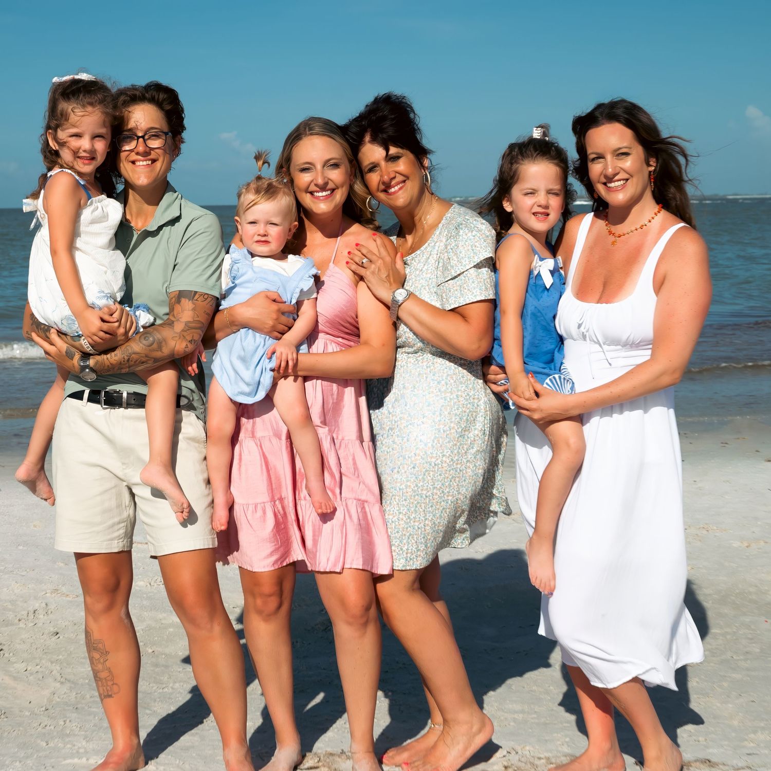 Family group smiling together on a sunny beach, with women holding young children, wearing summer outfits against a blue sky.