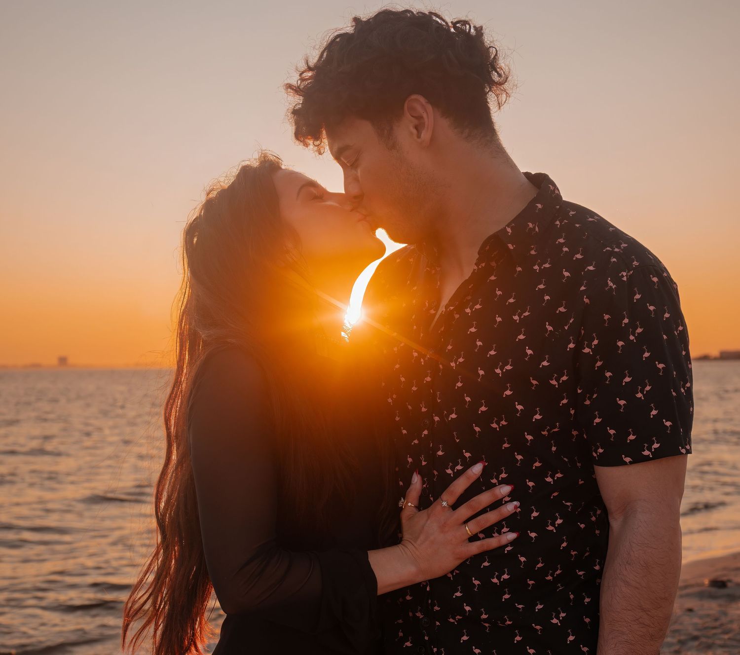 Young couple kissing at sunset by the ocean with warm golden light glowing between them.