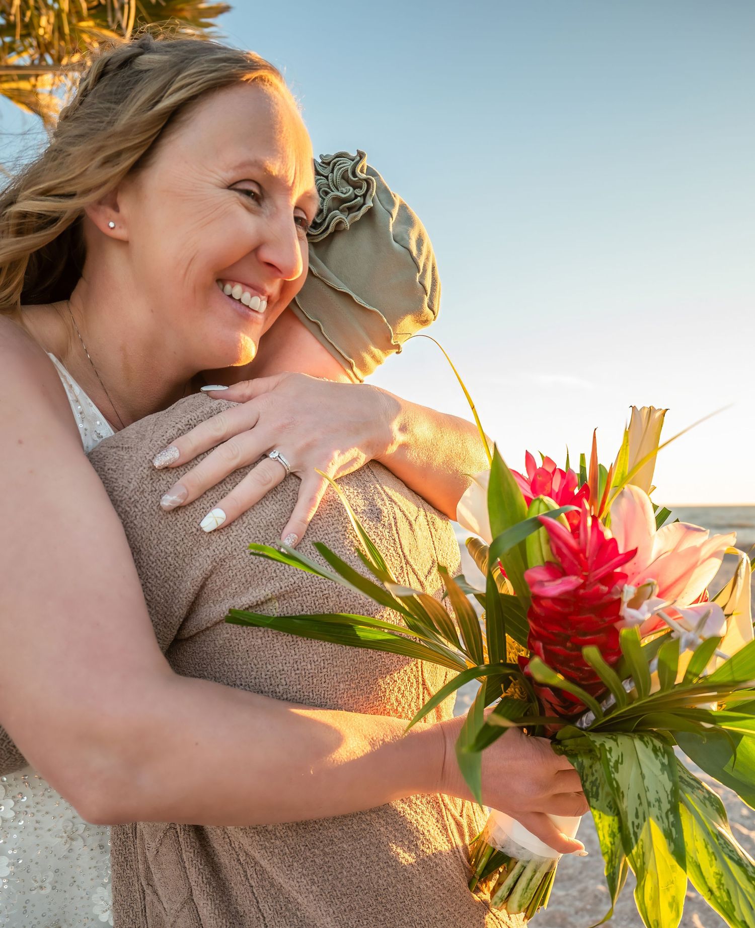 Joyful bride hugging a guest on the beach at sunset while holding a tropical bouquet.