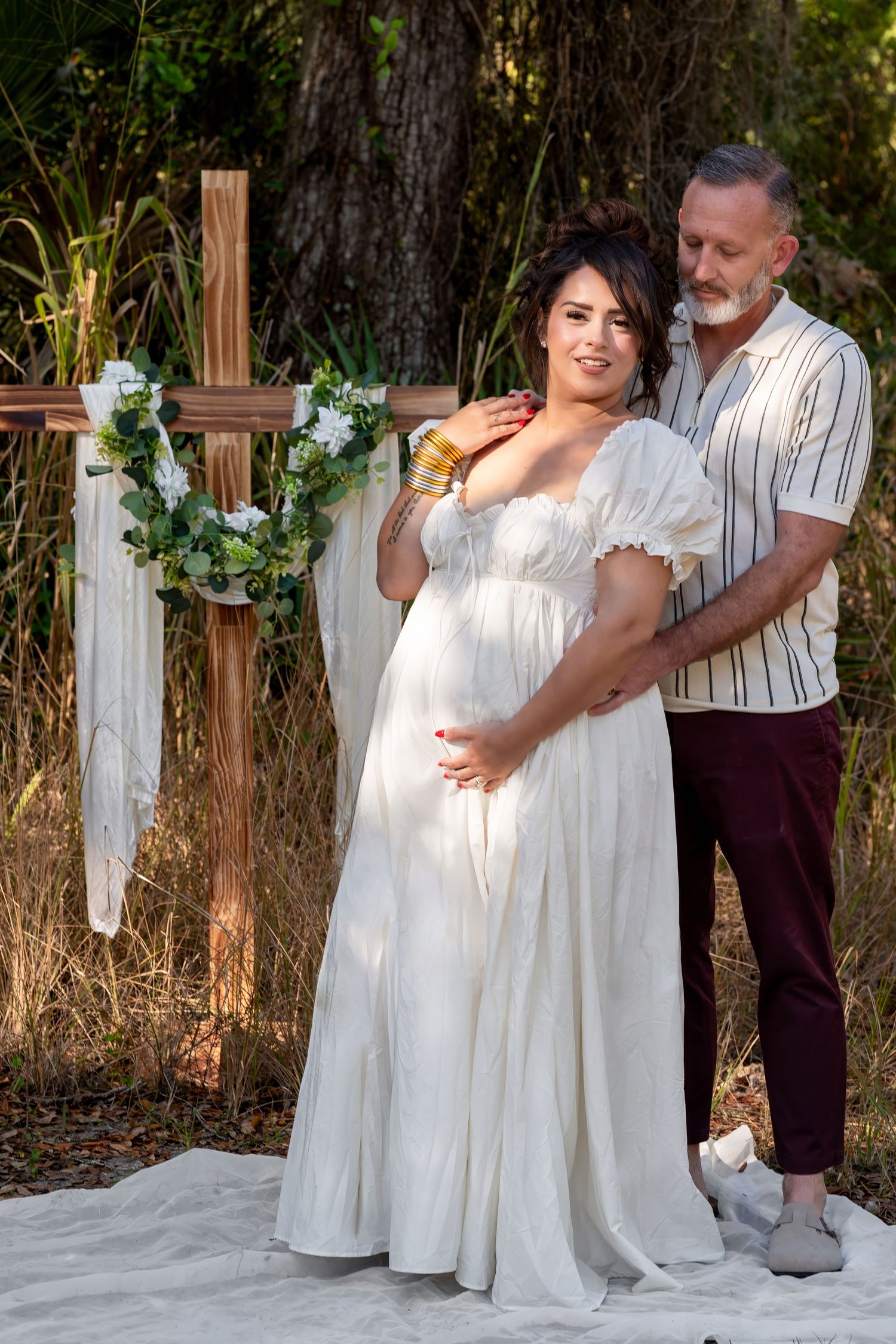 Couple posing outdoors near wooden cross, pregnant woman in white gown embraced by bearded man.