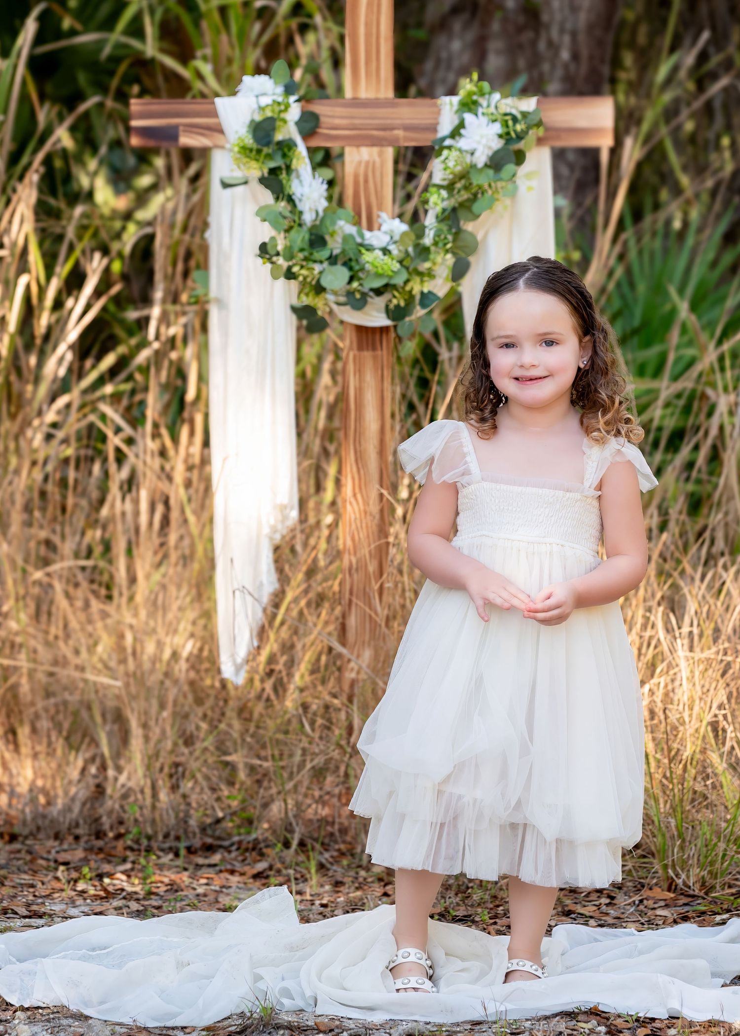 Adorable young girl in cream tulle dress standing in front of a wooden cross with floral wreath outdoors.