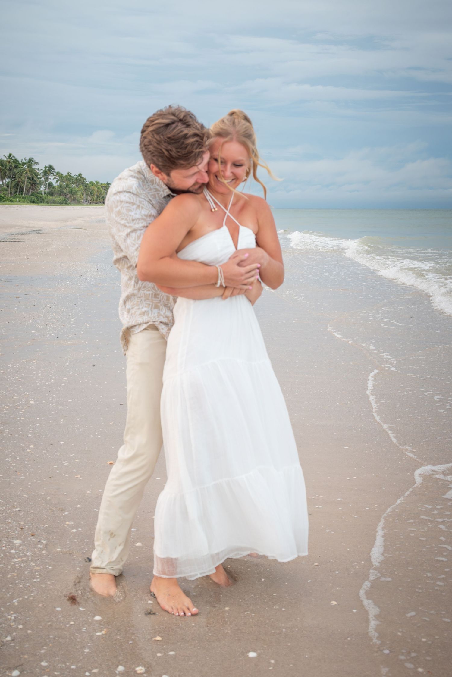 Groom embracing bride in white dress on sandy beach with ocean waves during romantic beach wedding.