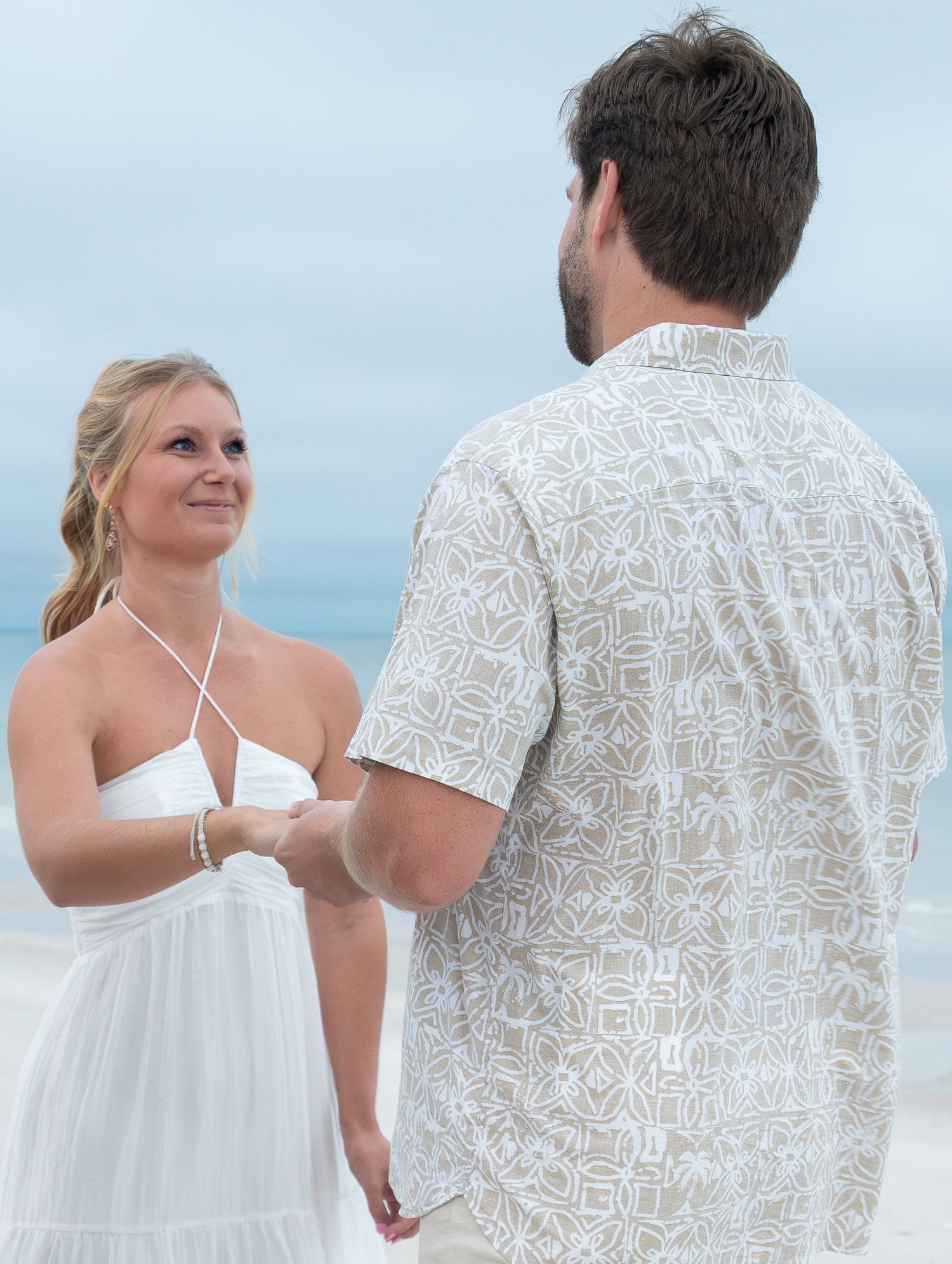 Couple holding hands during a romantic beach wedding ceremony, bride in white halter dress, groom in patterned shirt.