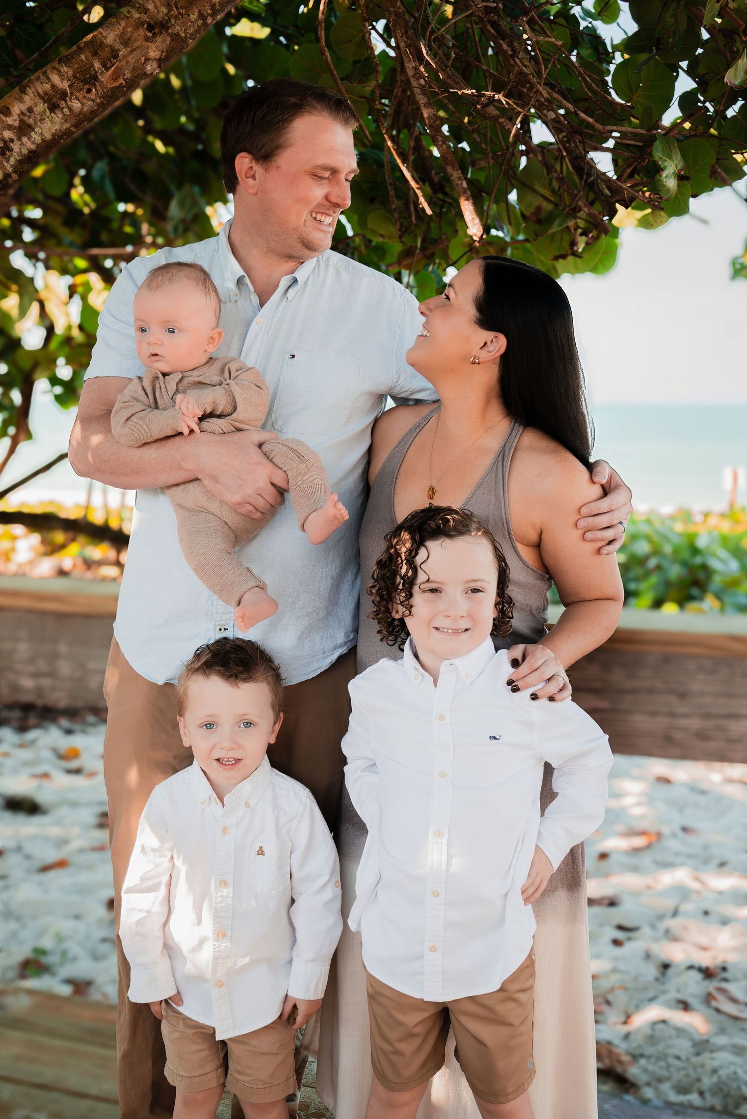 Happy family of five with baby and two boys in white shirts posing under a tree at a beach resort.