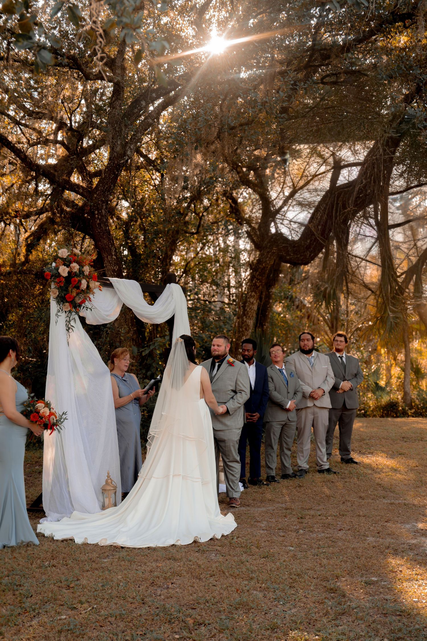 Sunlit outdoor wedding ceremony with bride, groom, officiant, and groomsmen beneath a draped floral arch.