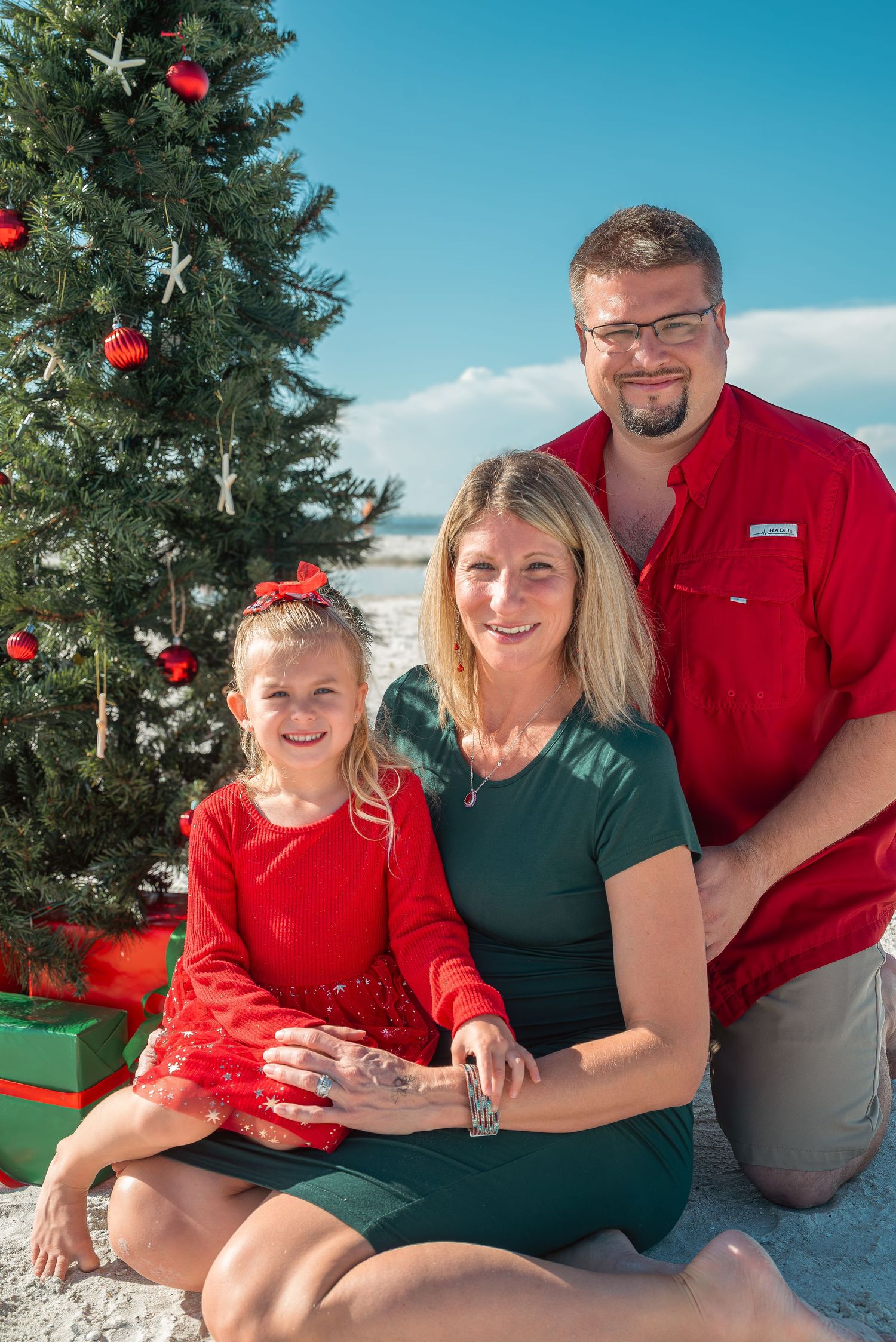 Family of three poses by decorated Christmas tree on sandy beach, pregnant mother in green dress with daughter and father.