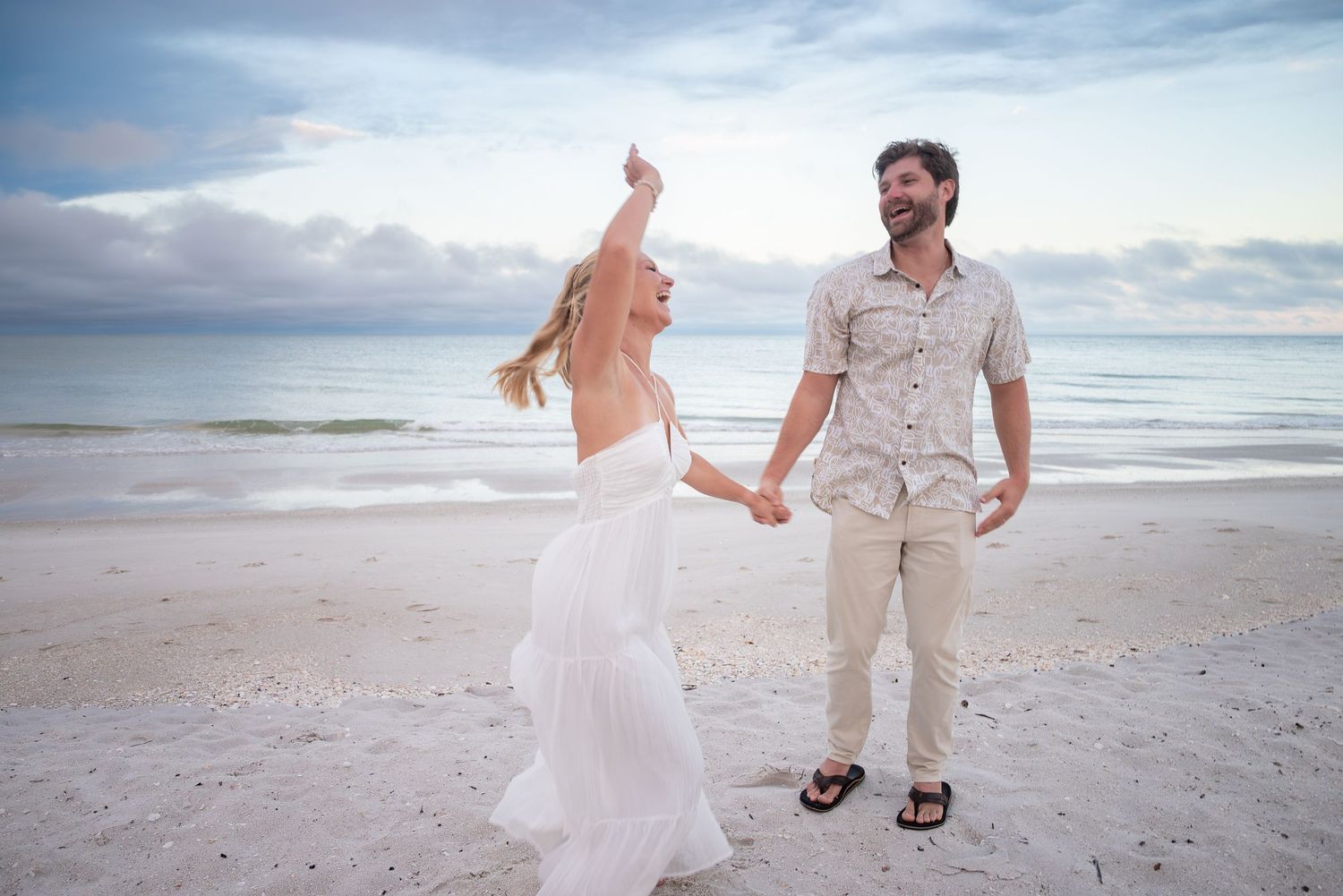 Couple dancing joyfully on a sandy beach at sunset, woman in white dress twirling, man in casual linen outfit.