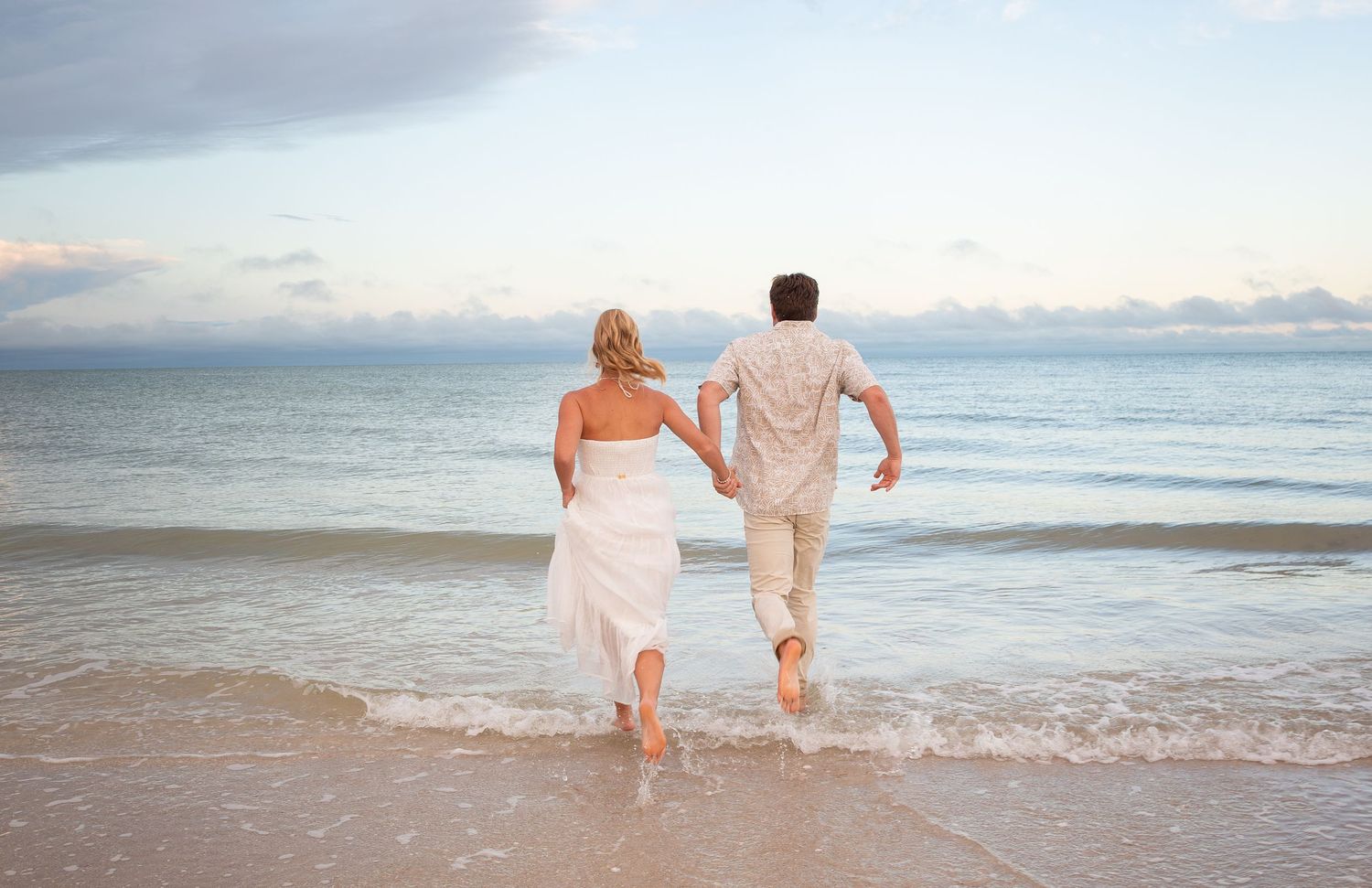 Couple in wedding attire running hand-in-hand along a sandy beach shore with ocean waves at sunset.
