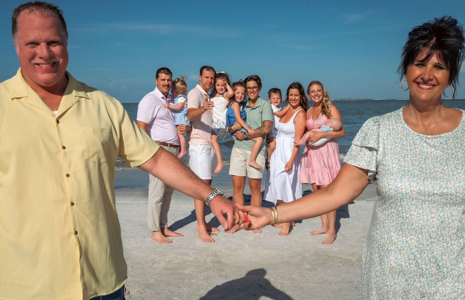 Large family holding hands on a sunny beach, smiling together with clear blue sky and calm ocean in the background.