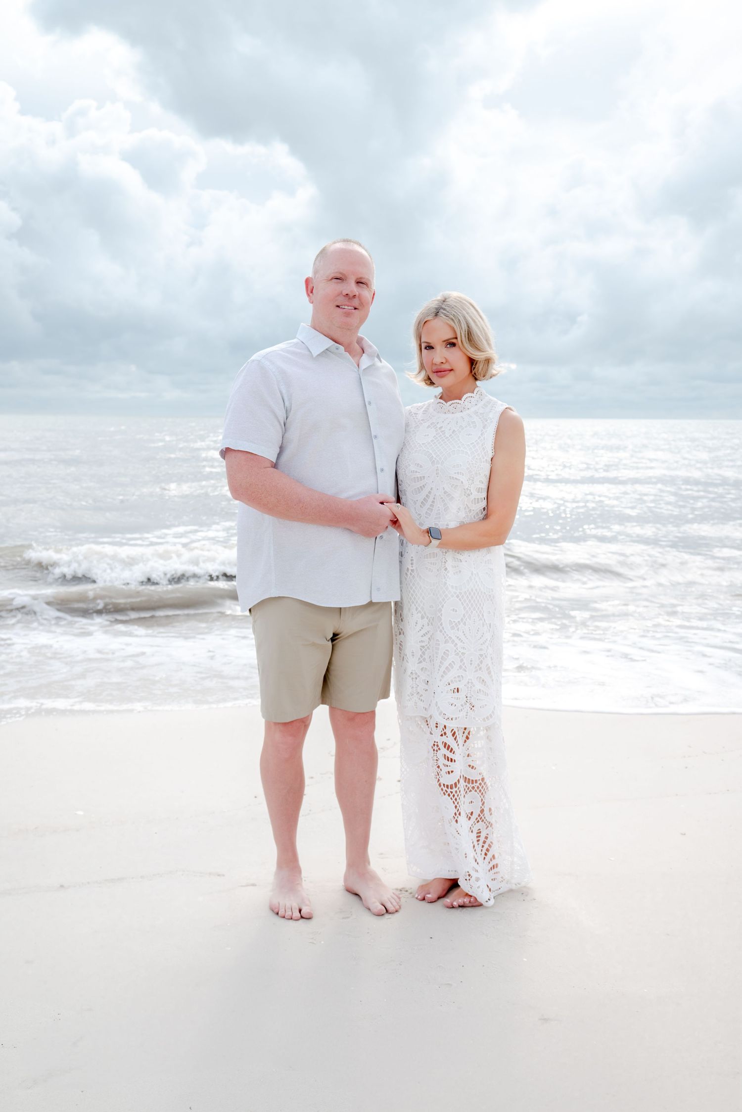 Couple posing together on sandy beach with ocean waves and cloudy sky in background.