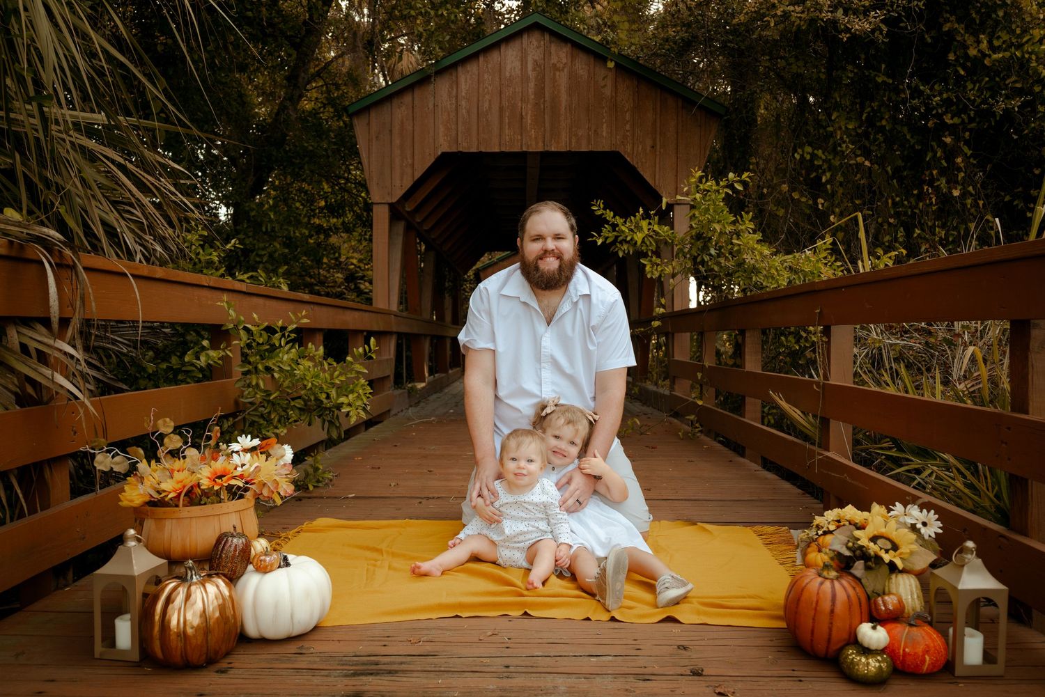 Father posing with two young children on wooden bridge with fall pumpkin decorations.