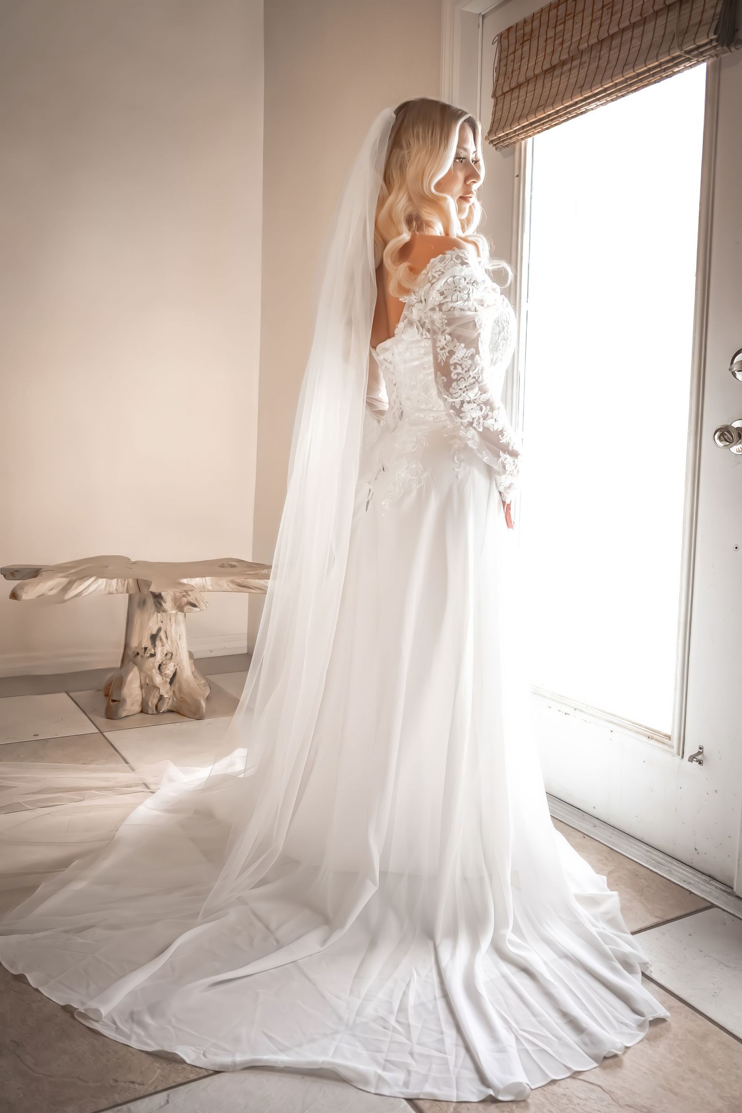 Elegant bride in off-shoulder lace wedding gown with long cathedral veil standing by window.