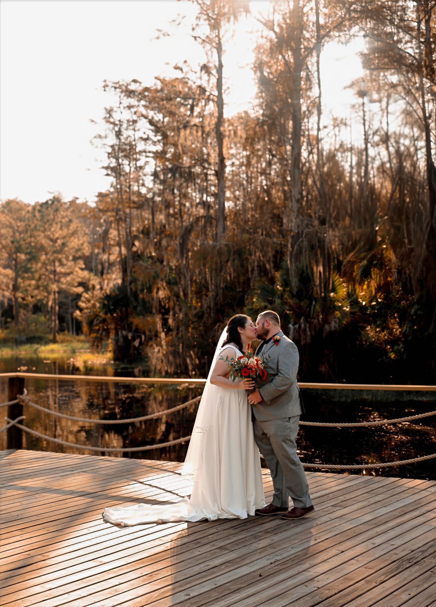 Bride and groom share a first kiss on a sunlit wooden dock overlooking a serene Florida swamp at sunset.