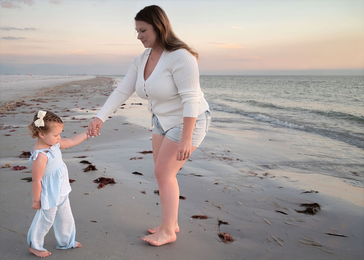 Mother and toddler daughter in white outfits holding hands and dancing on a peaceful beach at dusk.