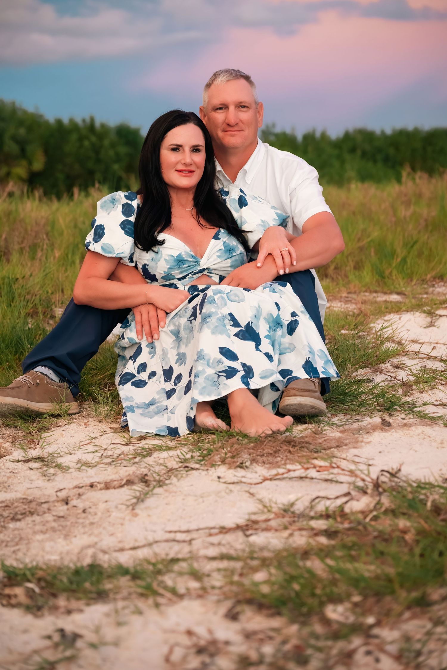 Couple sitting together on sandy ground in a grassy field, woman in floral dress, man in white shirt, at sunset.
