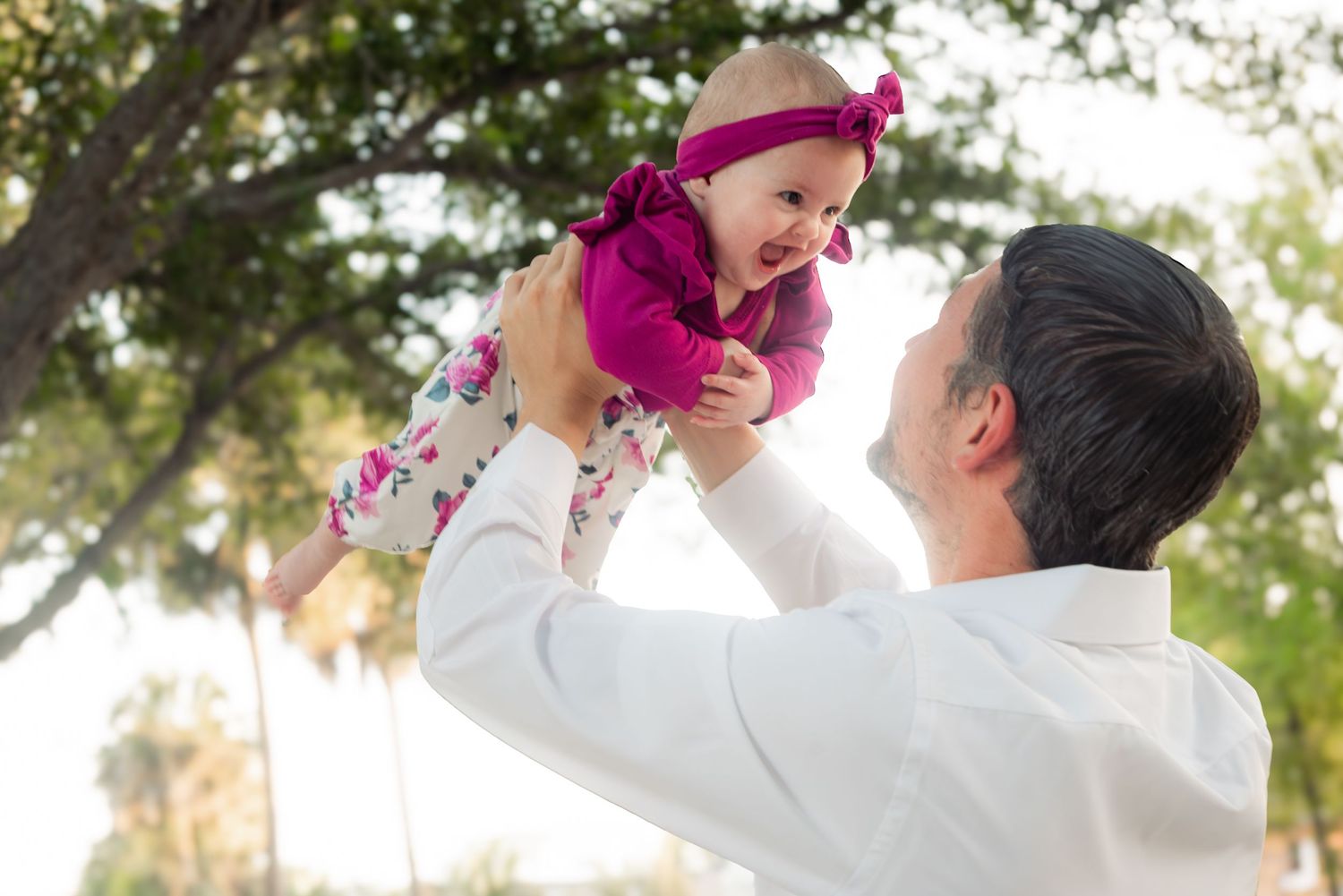 Loving father lifting his smiling baby girl dressed in pink up in the air outdoors near green trees.