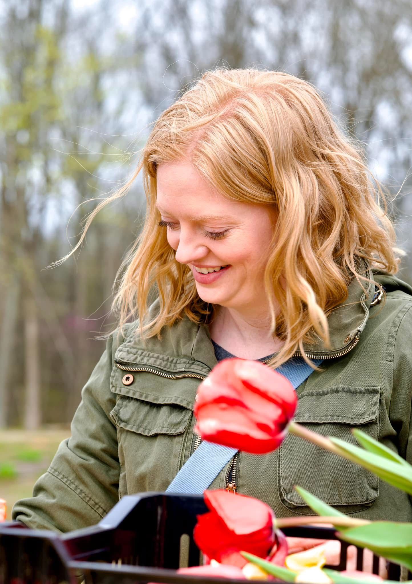 Happy blonde woman smiling while looking at red tulips in a basket outdoors.
