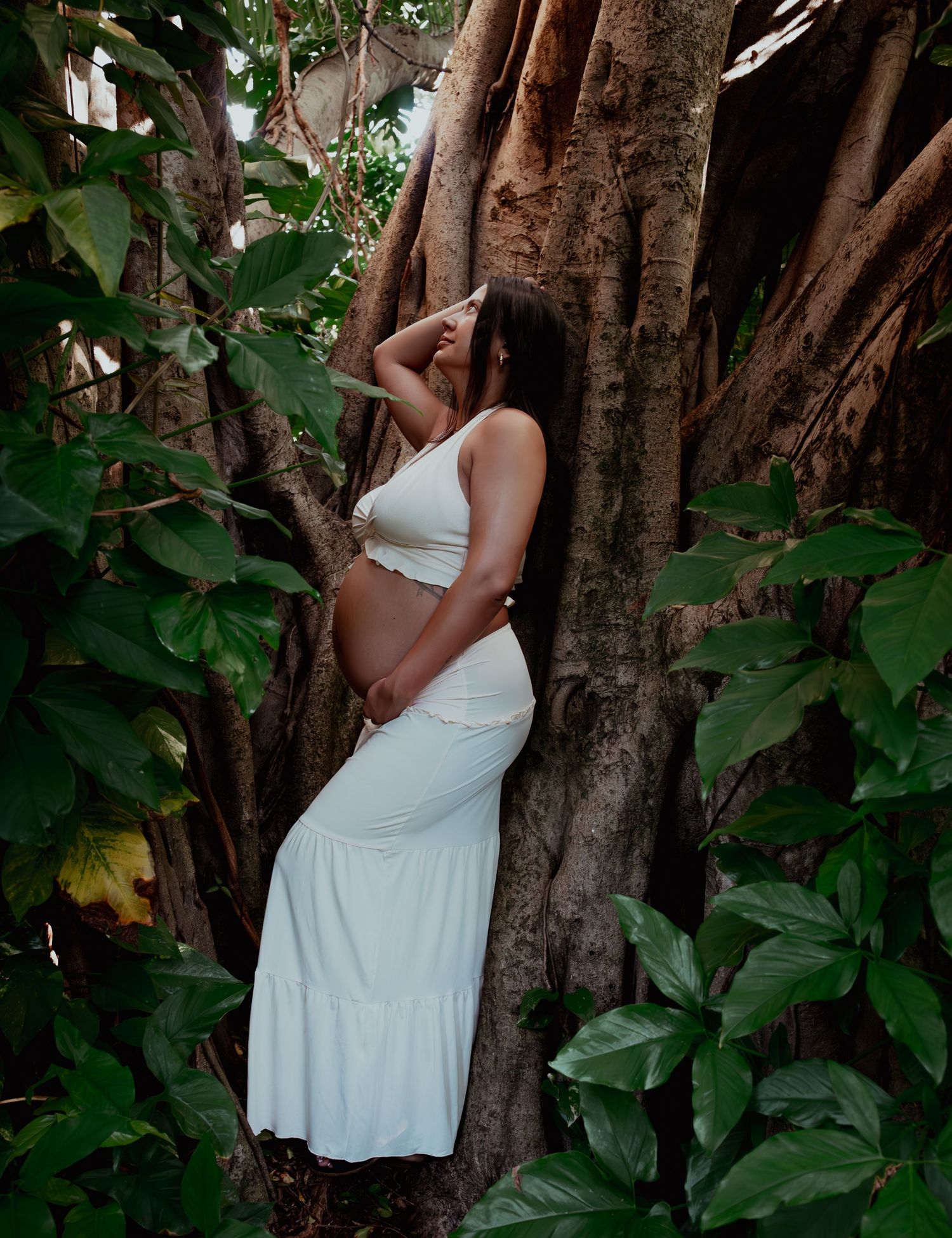 Pregnant woman in white outfit leaning against a large tree surrounded by tropical green foliage.