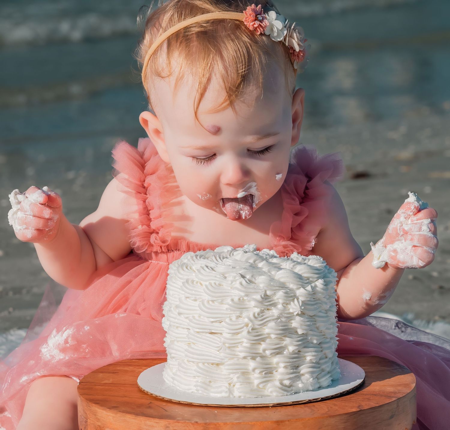 Baby girl in pink ruffle dress leaning into white birthday cake at beach.